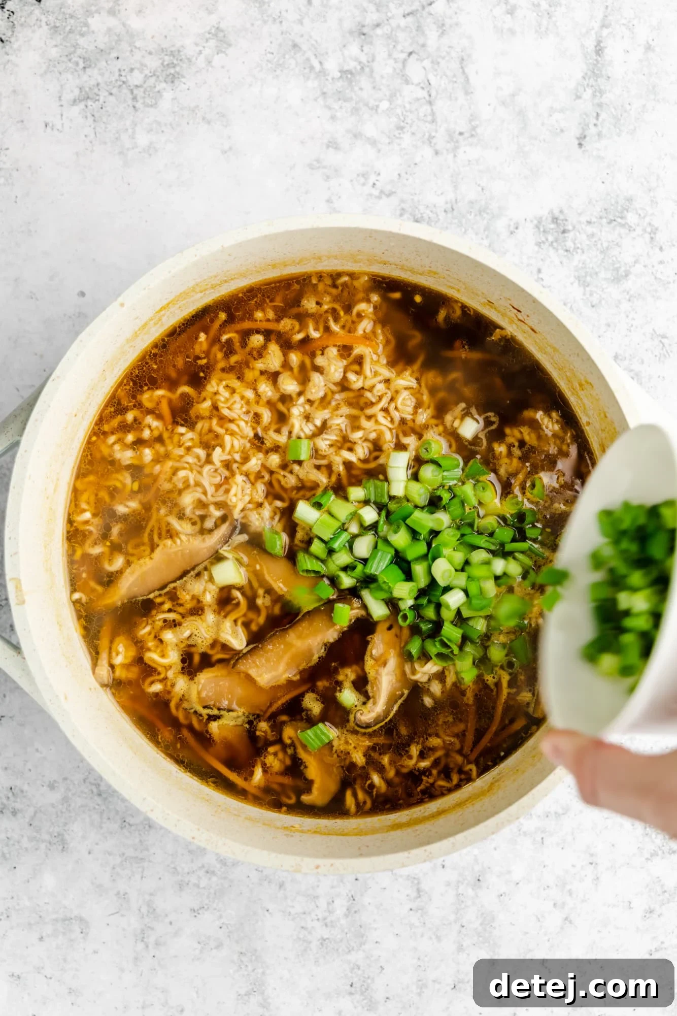 A hand pouring fresh diced scallions into a large pot of finished vegan ramen, adding a final touch of freshness and color.