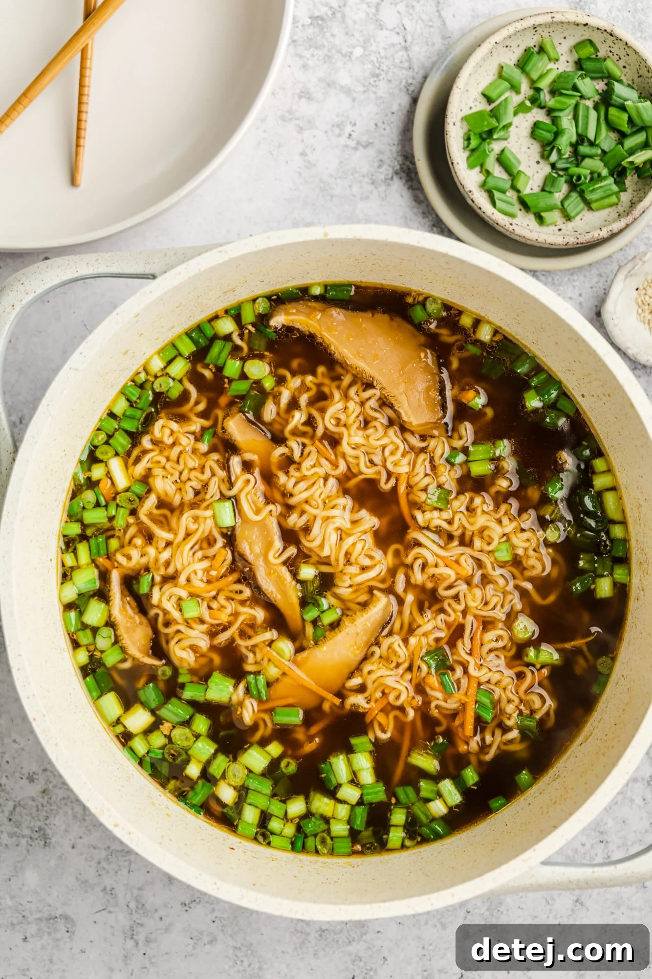 A large pot of vegan ramen with chopsticks leaning against the rim, next to a small bowl filled with freshly chopped scallions, highlighting the ease of adding toppings.