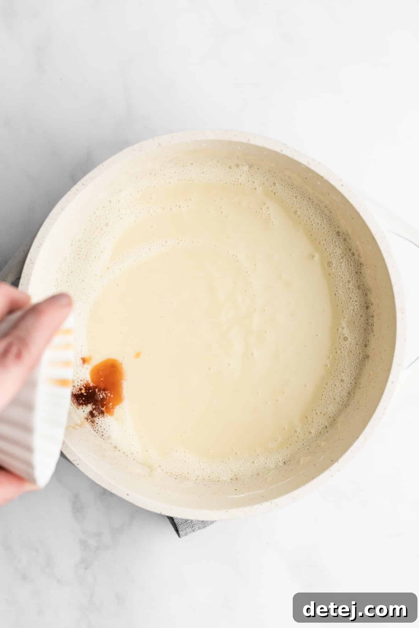 A woman's hand pouring vanilla extract into a creamy white custard in a saucepan.