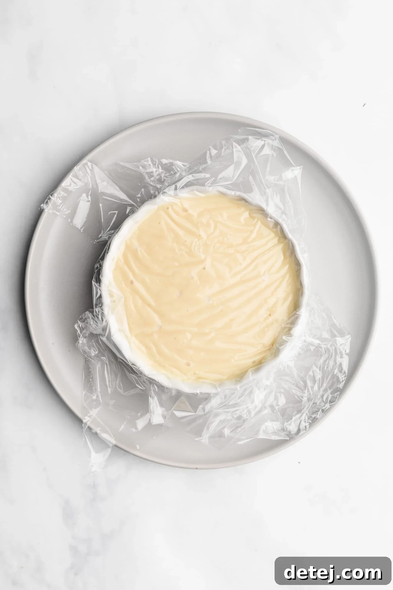 Vegan Creme Brulee in a white ramekin topped with a layer of plastic wrap, ready for chilling.