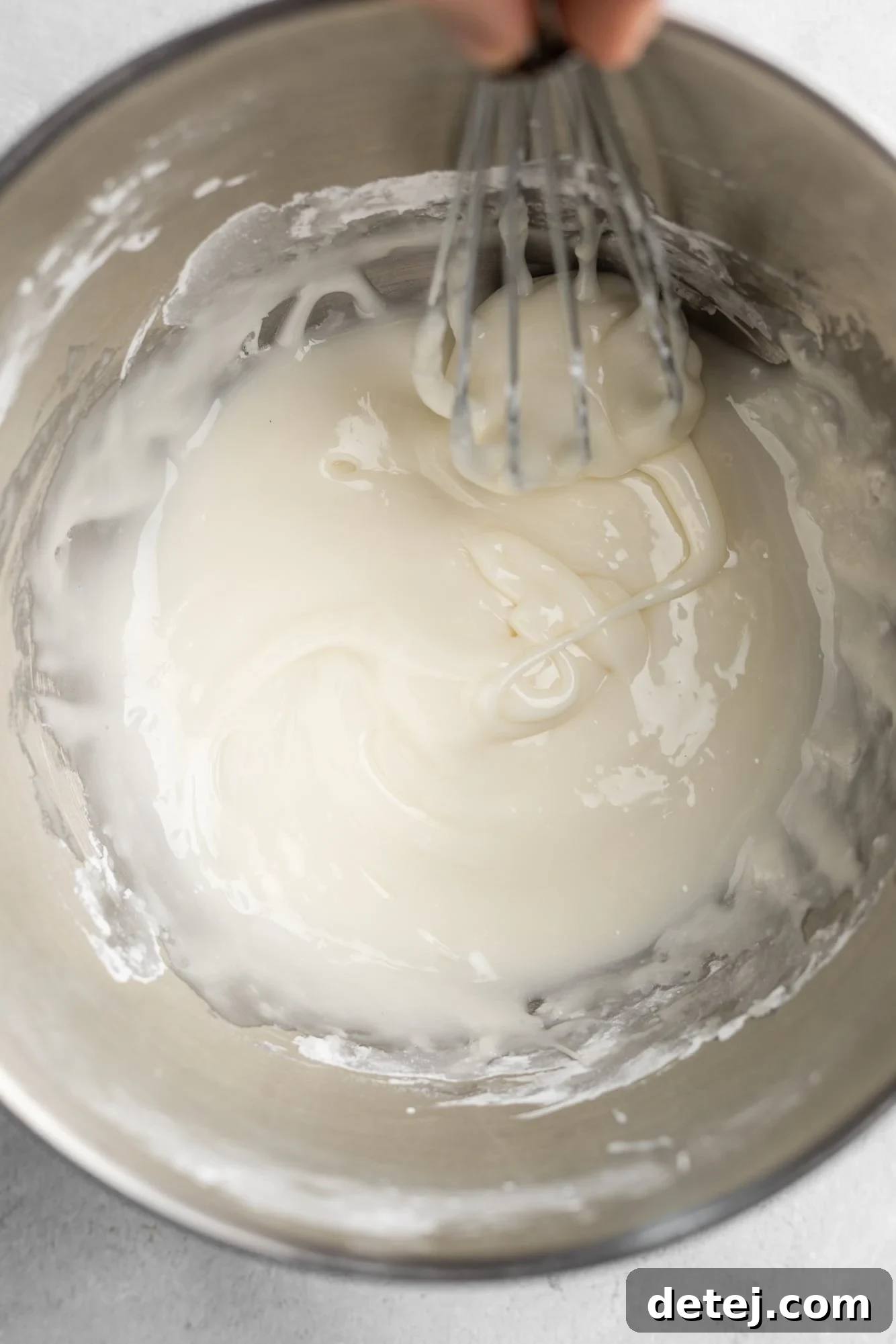 using a whisk to stir white icing in a large metal bowl.