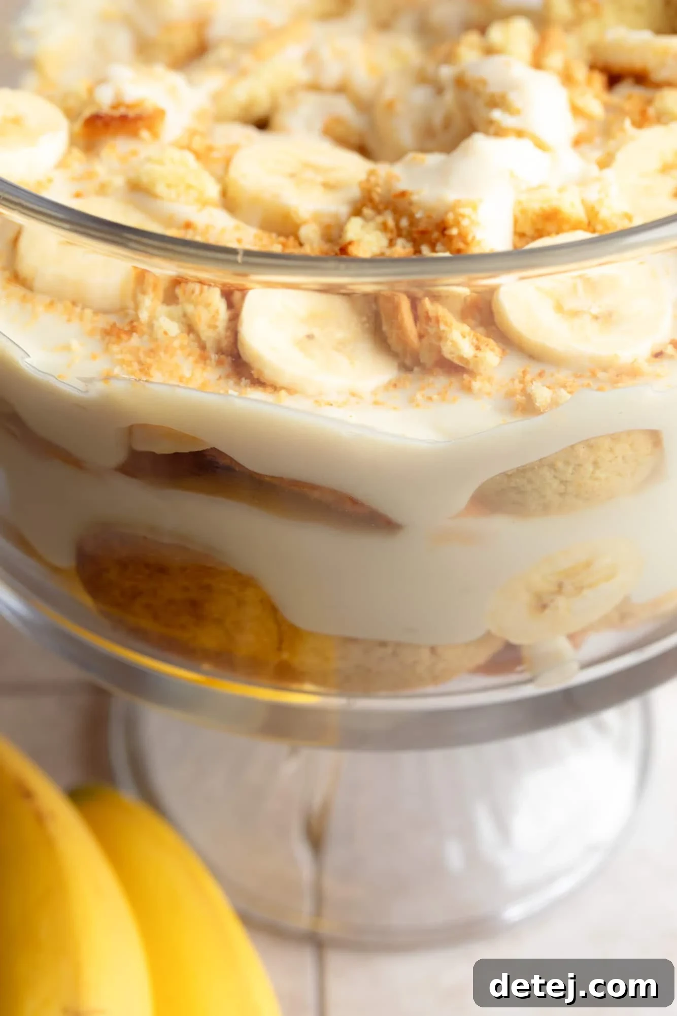 A clear glass trifle dish beautifully layered with golden vanilla custard, crisp vegan vanilla wafers, and neatly sliced bananas, ready to be chilled.