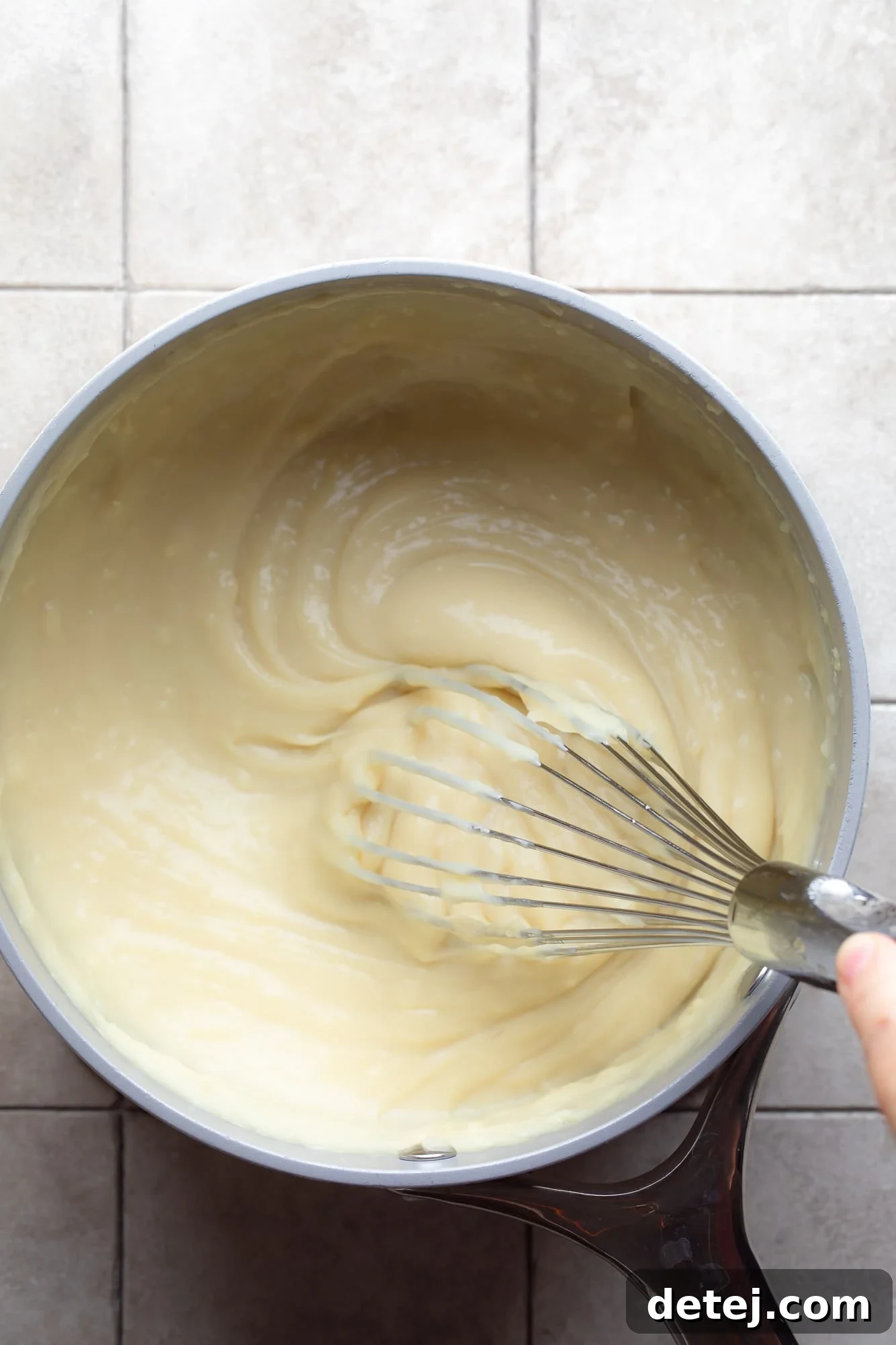 A close-up shot of a whisk actively stirring a creamy, pale yellow custard in a sleek grey pot, emphasizing the smooth consistency.