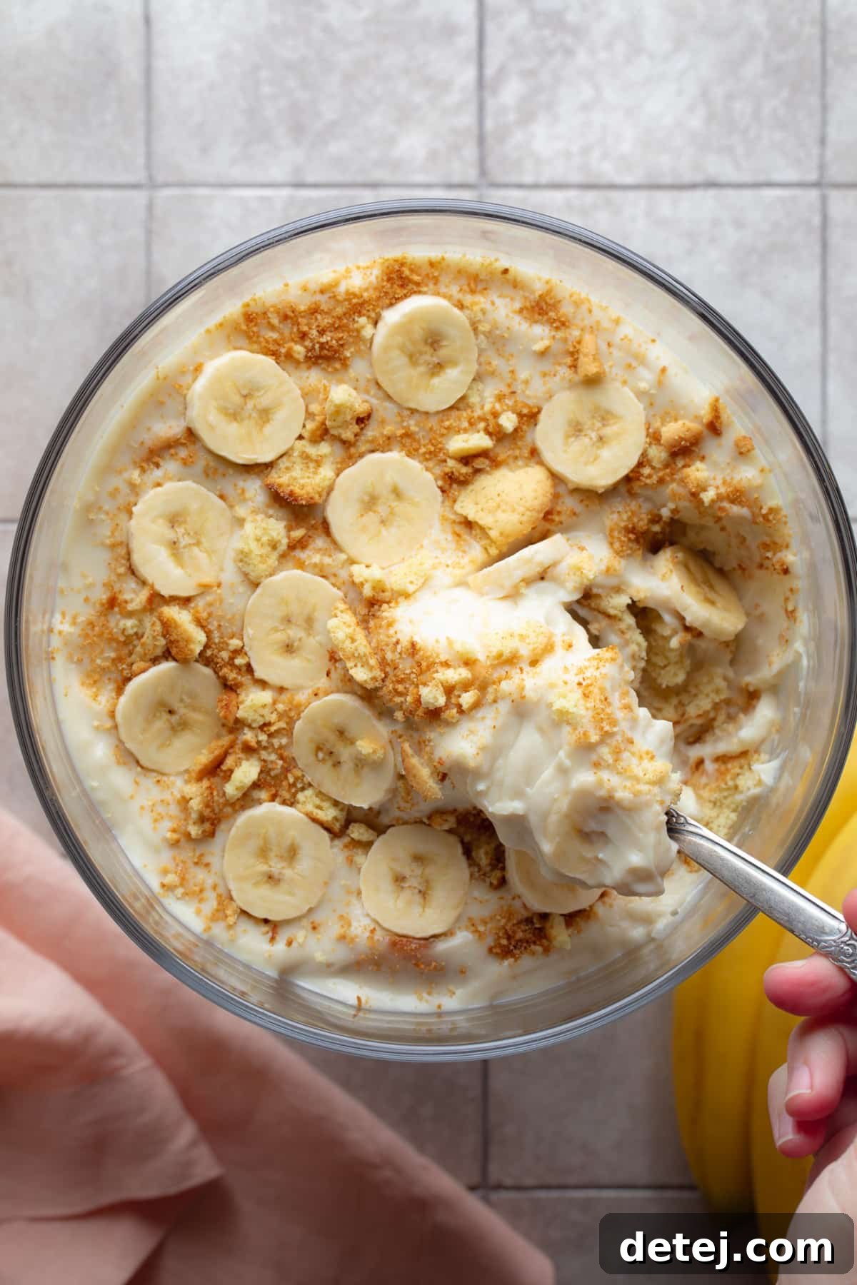 A large, clear glass dish filled to the brim with perfectly layered vegan banana pudding, showing delicious interplay of banana slices, rich cream, and delicate cookie crumbles against a grey background.