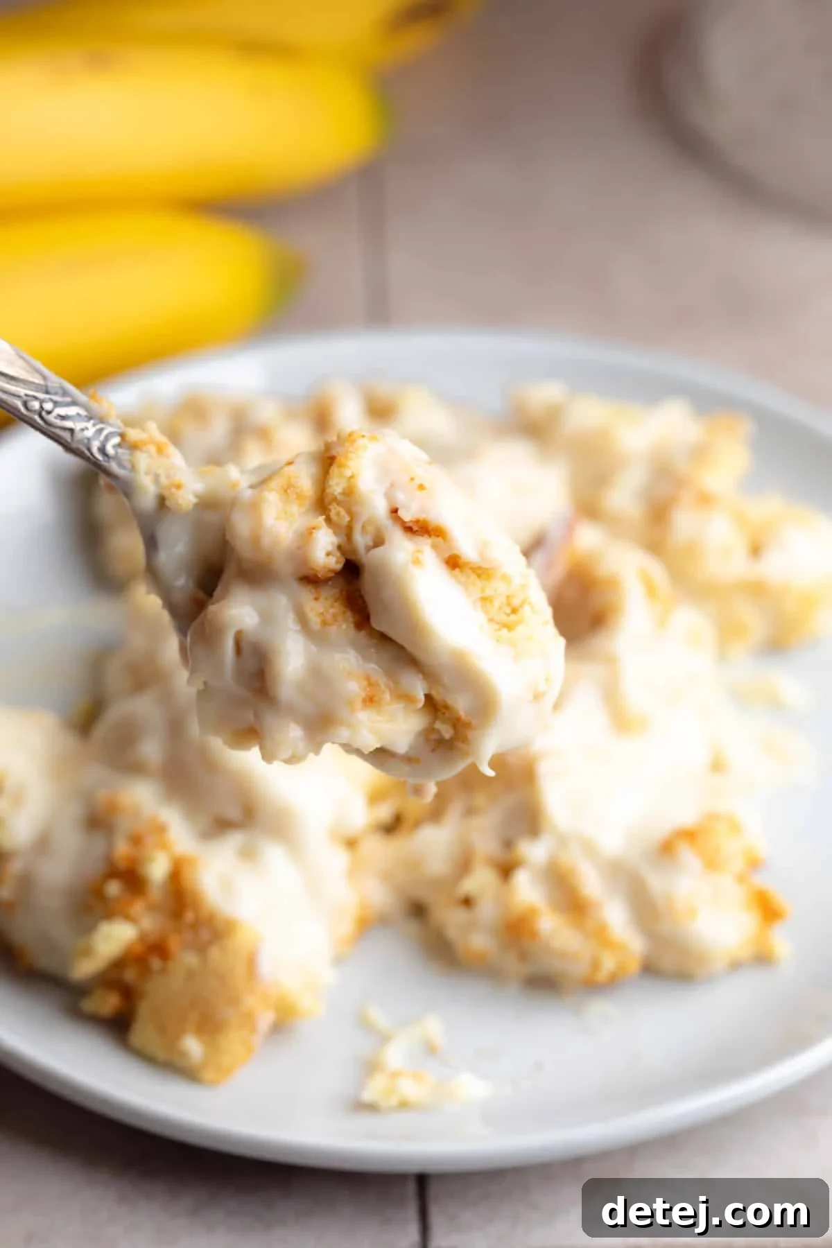 A close-up of a plate featuring a portion of creamy vegan banana pudding, with a spoon taking a bite, showing the layers of custard, cookies, and bananas.