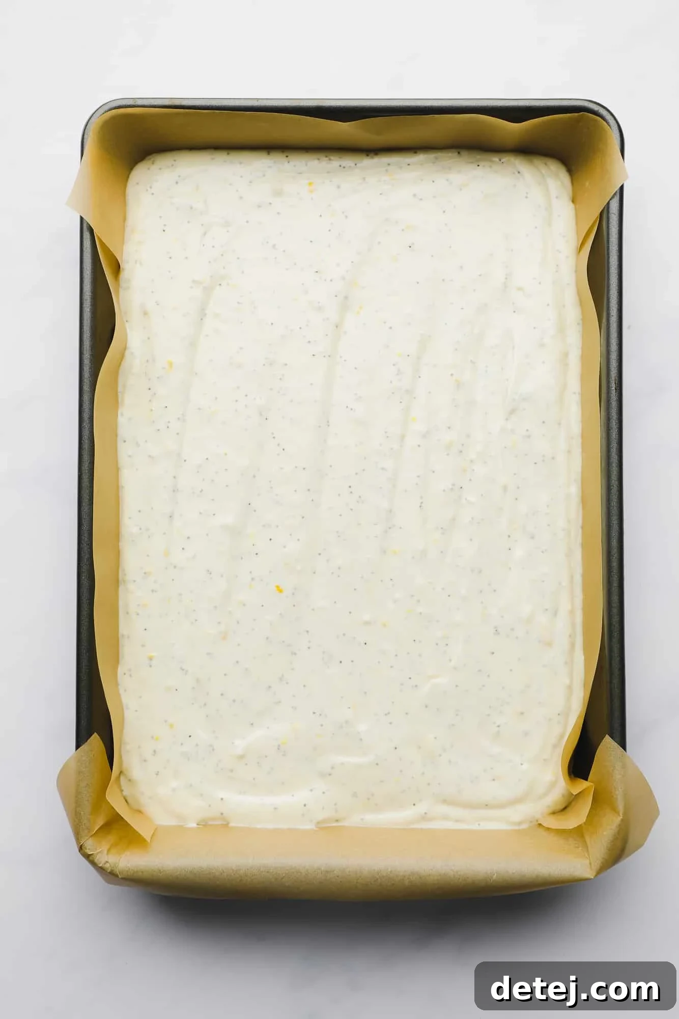 Smooth, pale yellow vegan lemon poppyseed cake batter, studded with poppy seeds, resting evenly in a large rectangular baking pan, ready for the oven.