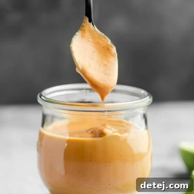 A spoon lifting up from a small glass jar of orange chipotle sauce.