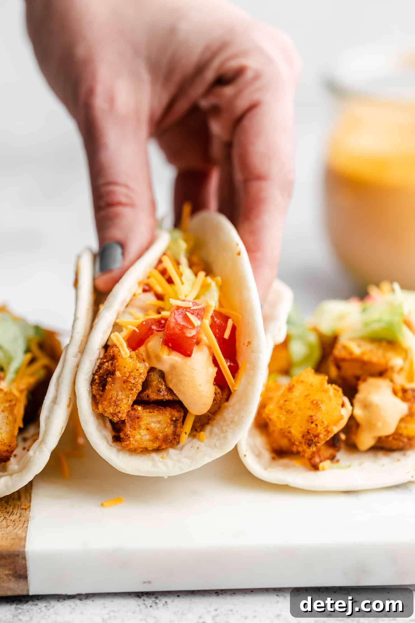 A woman's hand reaching for a vegan spicy potato taco from a row of several others, all perfectly assembled.