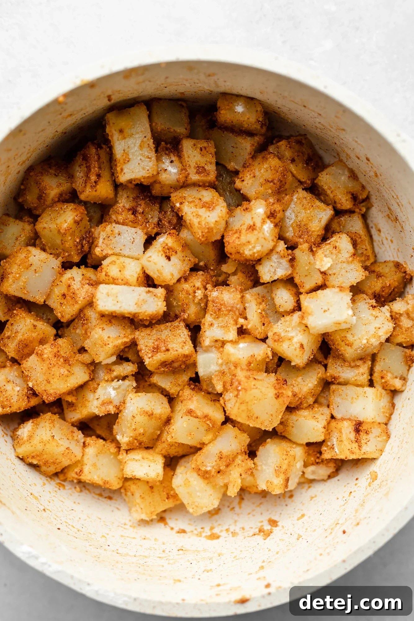 Parboiled and seasoned potato chunks, glistening with oil and spices, resting in a large white mixing bowl.