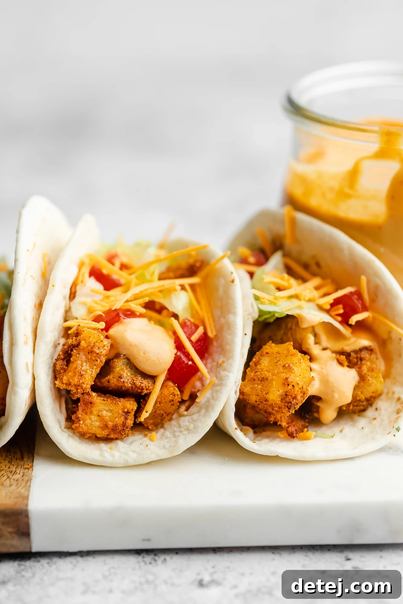 A close-up shot of two spicy potato tacos, generously topped with shredded vegan cheddar cheese, creamy chipotle sauce, fresh lettuce, and diced tomatoes, sitting side by side.