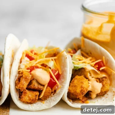 Close up on 2 potato tacos next to each other topped with shredded cheddar cheese, chipotle sauce, lettuce, and tomatoes.