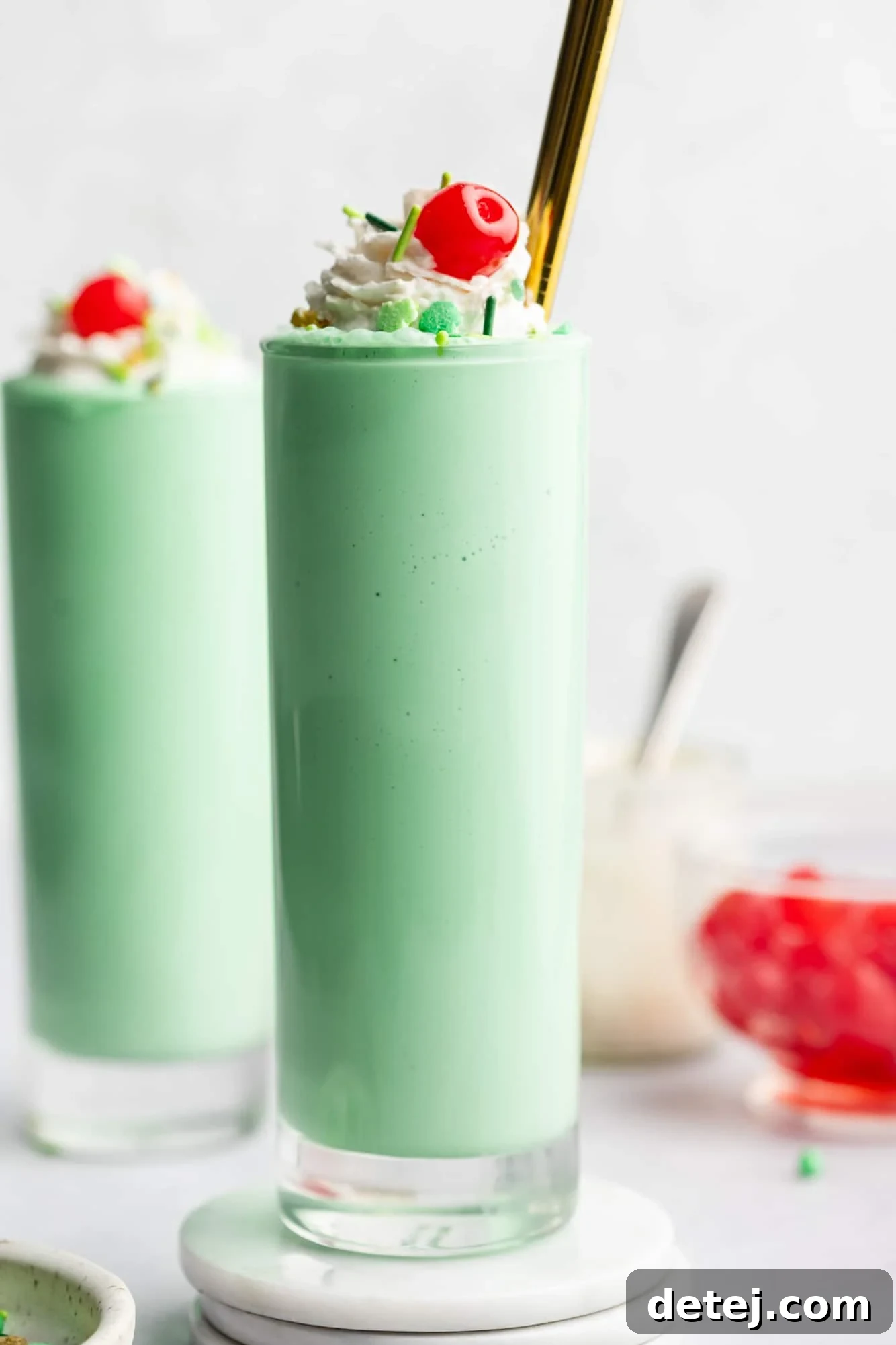 Two vibrant mint green vegan shamrock milkshakes in tall glasses, generously topped with fluffy vegan whipped cream, festive green sprinkles, and a glistening maraschino cherry.