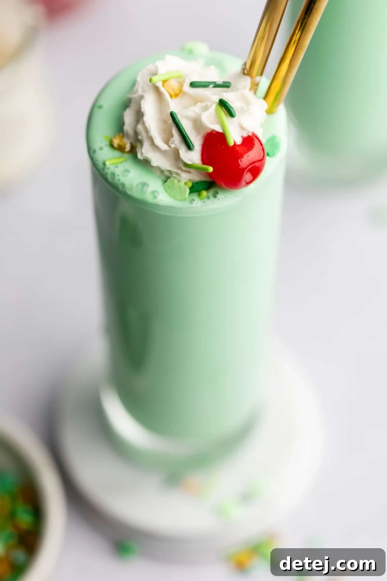 A close-up view of a single vegan shamrock shake in a tall, elegant glass, adorned with a generous dollop of vegan whipped cream, sparkling green sprinkles, and a bright red maraschino cherry.