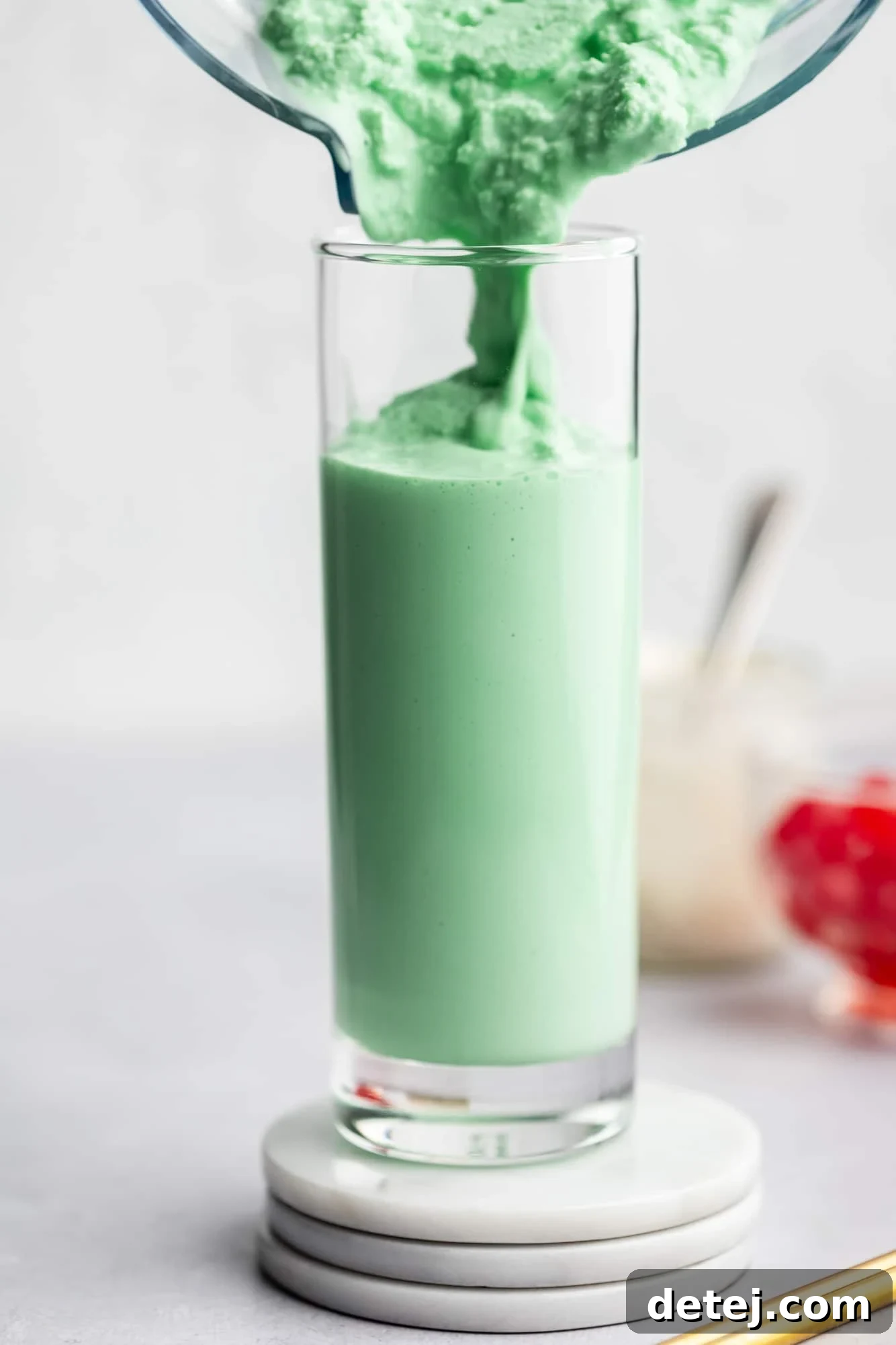 A stream of perfectly blended, mint green vegan shamrock shake being poured gracefully into a tall, clear glass, creating a sense of anticipation.