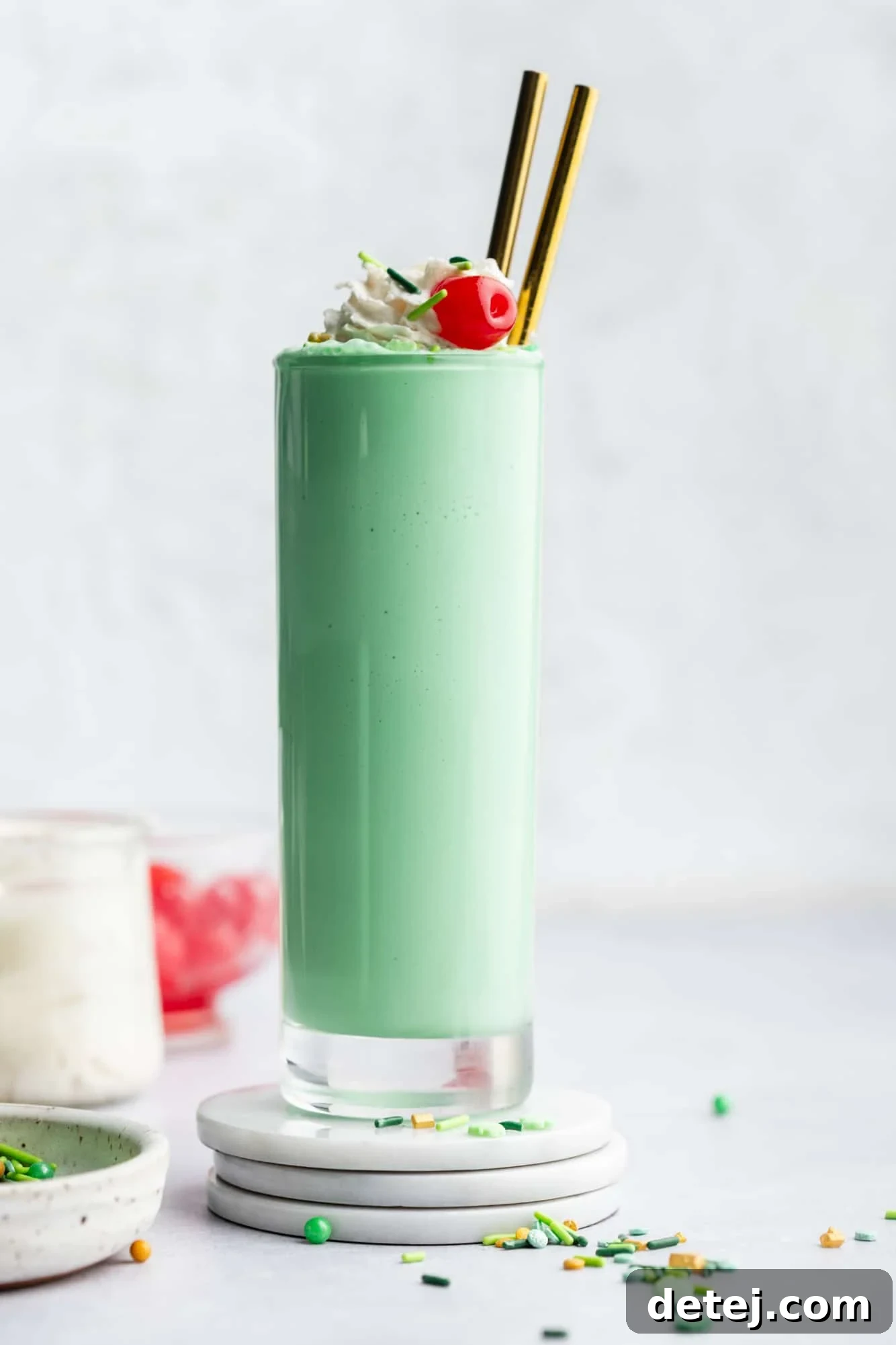 A single, perfectly presented vegan shamrock shake in a tall glass, beautifully topped with vegan whipped cream, sparkling green sprinkles, and a vibrant maraschino cherry.