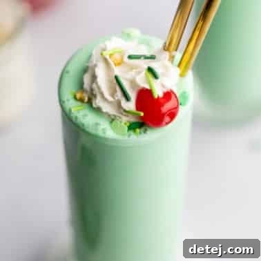 close up on a vegan shamrock shake in a tall glass topped with vegan whipped cream, green sprinkles, and a maraschino cherry.