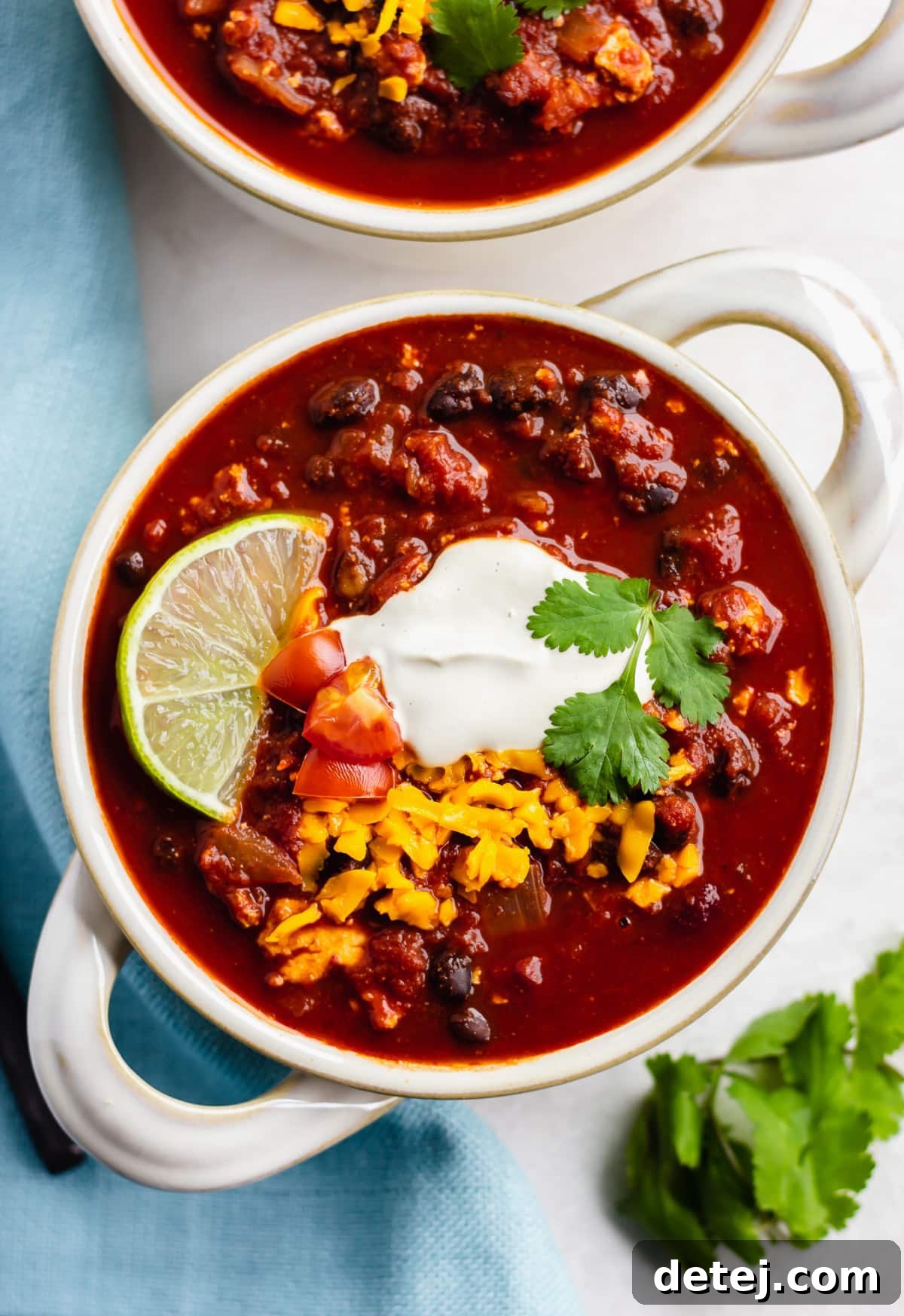 looking down on two bowls of chili with sour cream, cheese and a lime on white background with blue towel