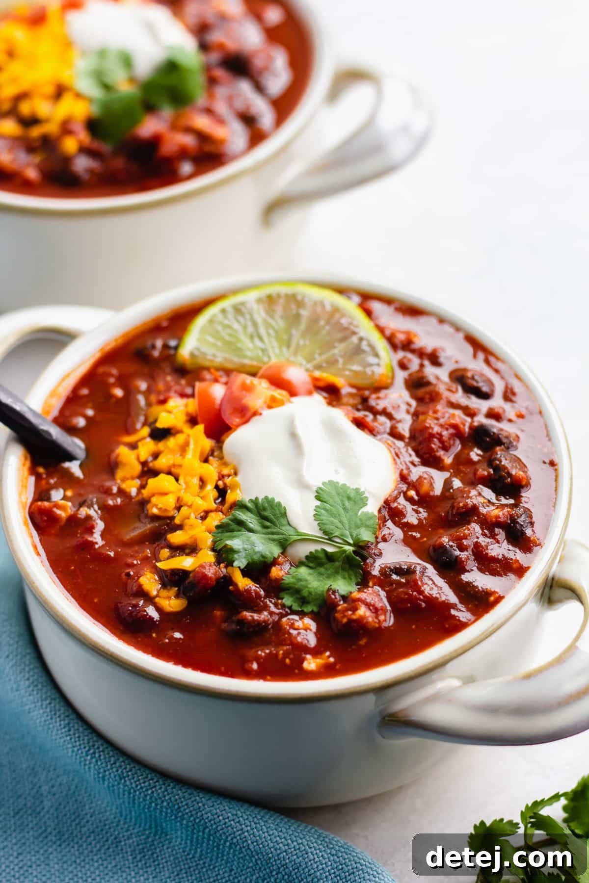 two bowls of vegan chili on white background with blue towel, sour cream and cheese on top.