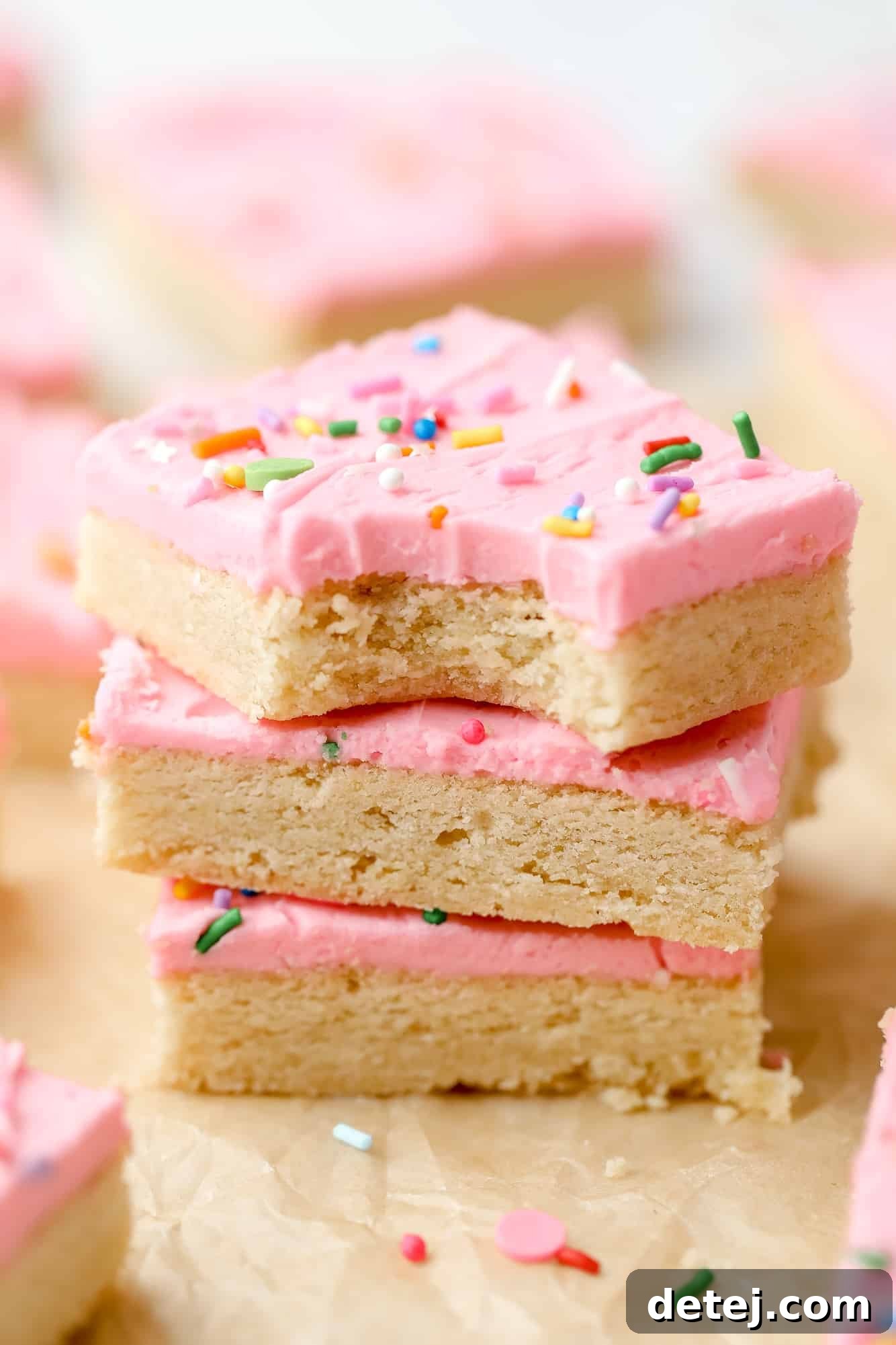 A stack of three vegan sugar cookie bars, each generously topped with fluffy pink frosting and colorful sprinkles.