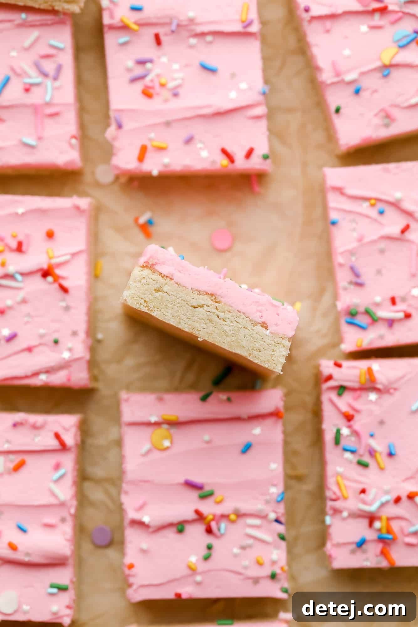 A single vegan sugar cookie bar, tipped on its side to show its thickness, rests beside rows of more pink-frosted bars, ready to be enjoyed.