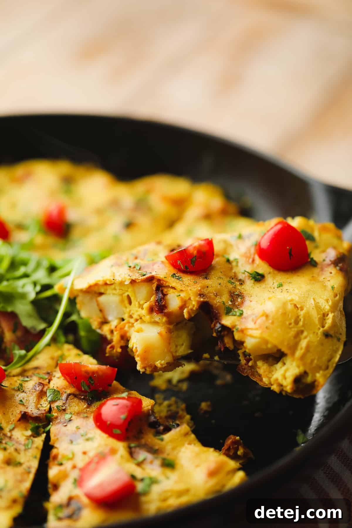 lifting a slice of vegan frittata topped with sliced cherry tomatoes from a black skillet.