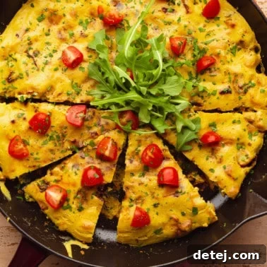 a cooked vegan frittata topped with sliced cherry tomatoes and arugula in a large black skillet.
