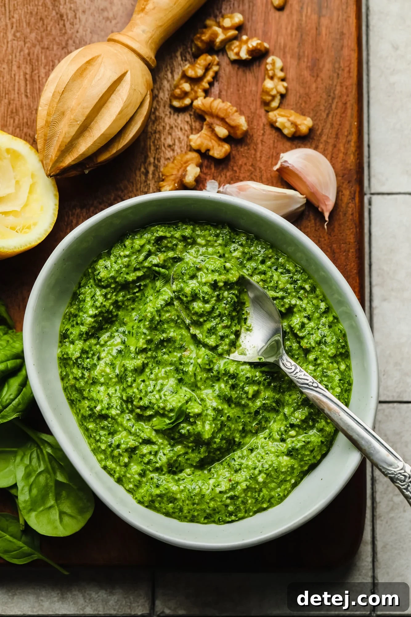 A beautiful grey bowl filled with vibrant green spinach pesto, with a spoon resting in it. Fresh ingredients like spinach leaves, walnuts, garlic cloves, and a lemon are artfully arranged around the bowl, highlighting the wholesome components of this homemade sauce.