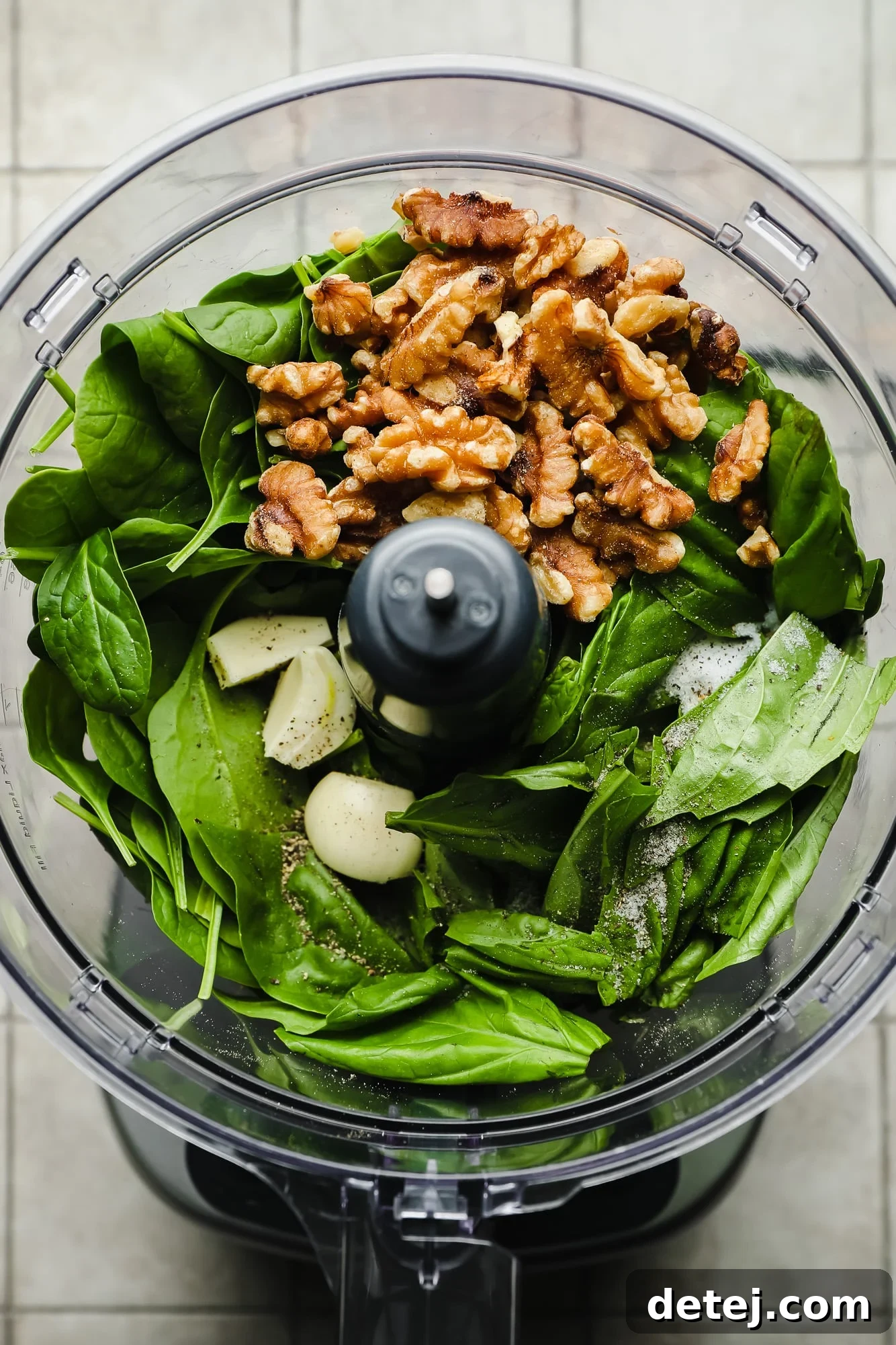 A food processor filled with fresh ingredients for spinach pesto, including vibrant spinach leaves, aromatic basil, garlic cloves, and walnuts, ready to be blended into a delicious sauce.