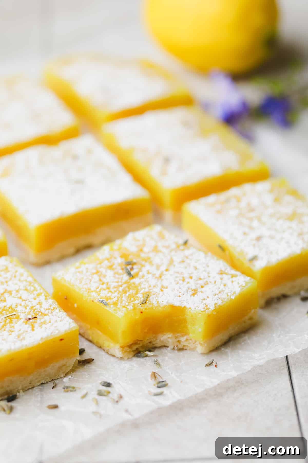 A perfectly portioned vegan lavender lemon bar with a bite taken out, revealing the creamy lemon filling, with more bars in the background on parchment paper adorned with delicate lavender flakes.