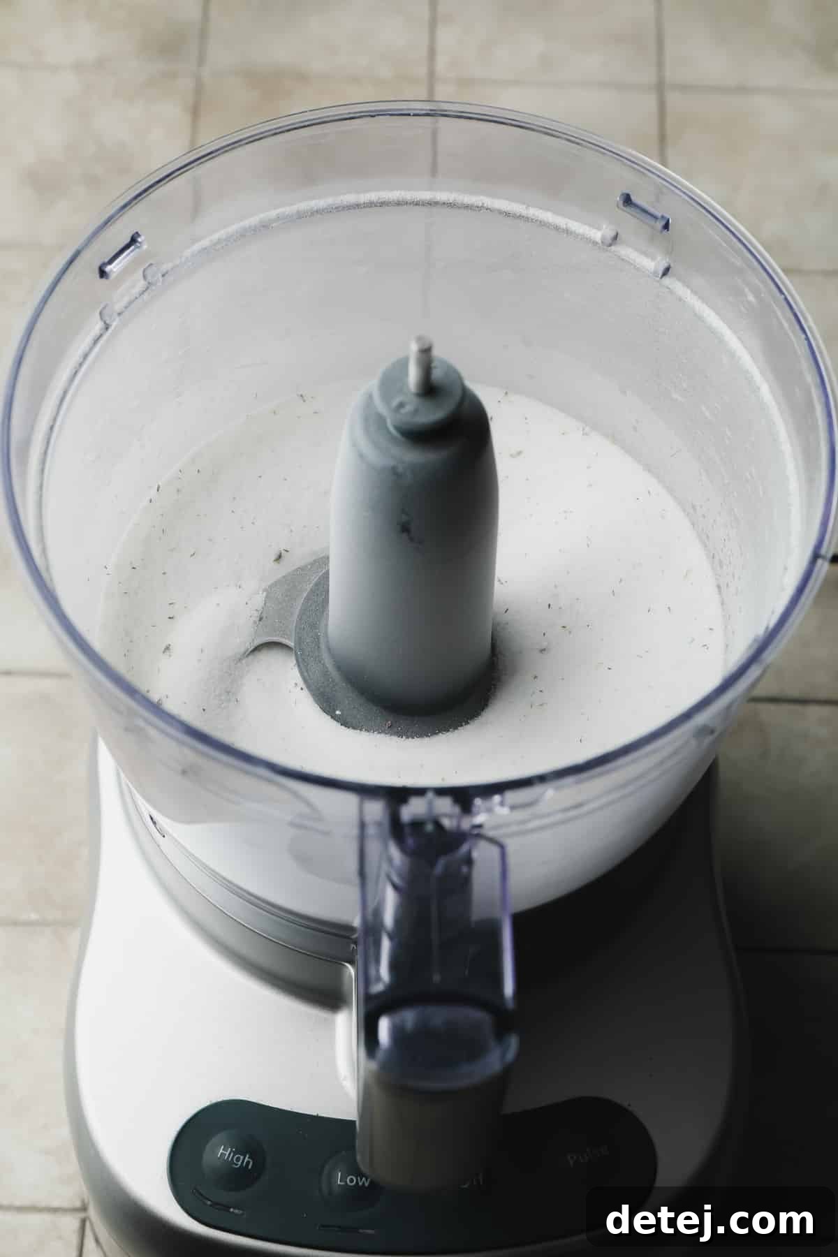 A food processor containing granulated sugar and small, dark specks of culinary lavender, ready to be blended into fragrant lavender sugar, set against a clean grey background.