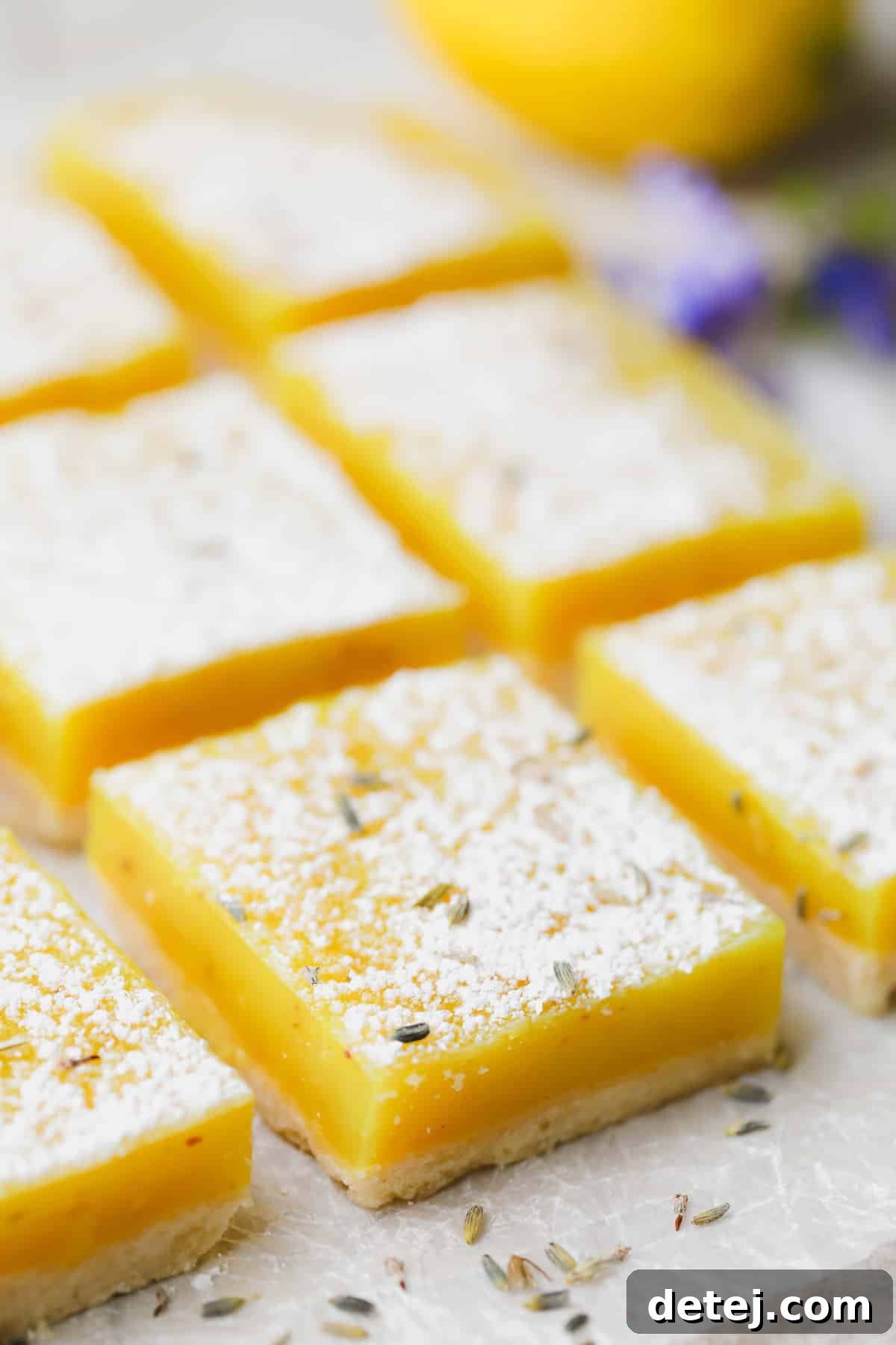 A side view of several neatly sliced vegan lavender lemon squares, with fresh lemon slices and delicate purple flowers in the softly blurred background, highlighting their elegant appeal.