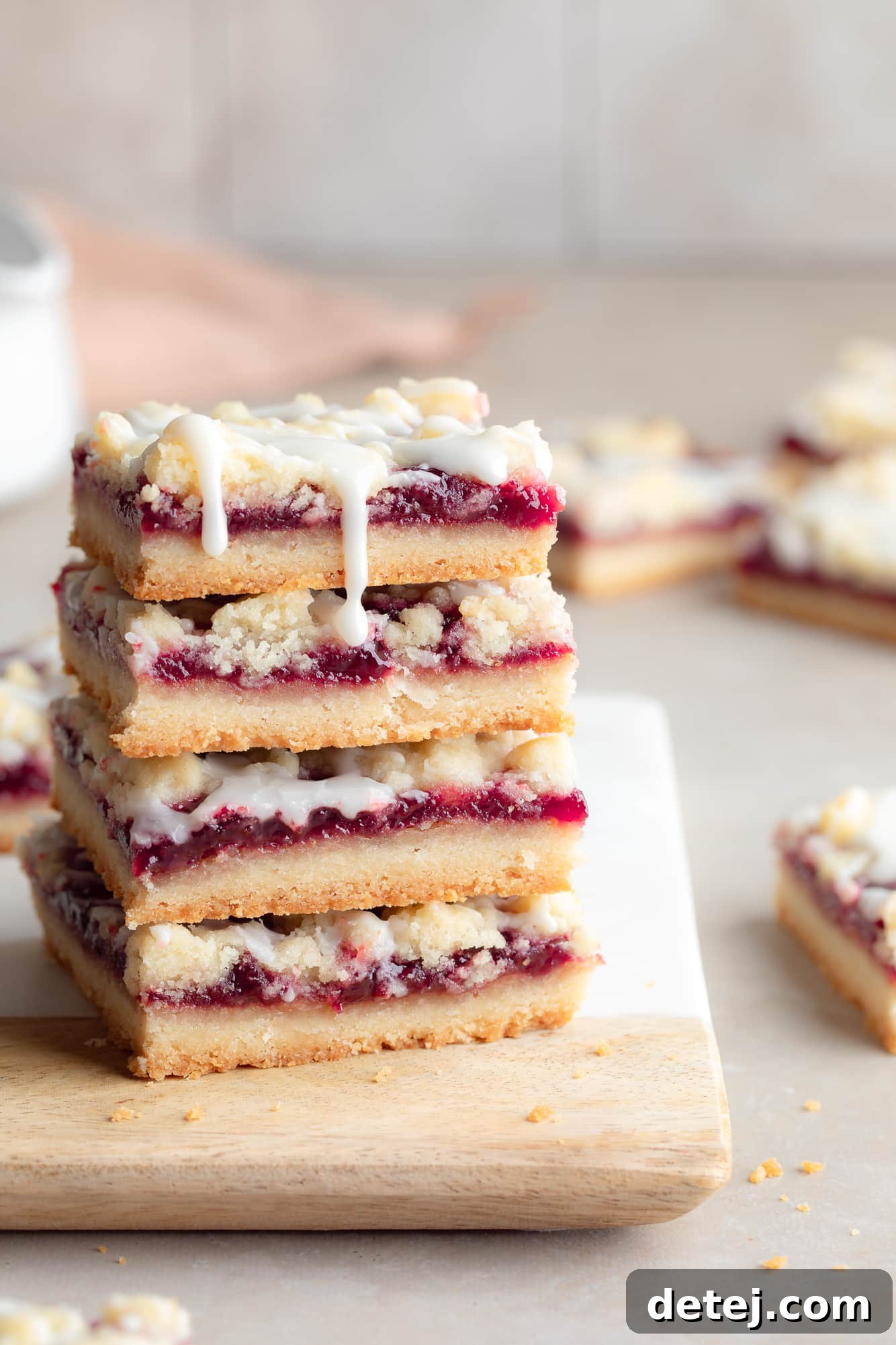 A close-up view of a stack of four perfectly golden-brown vegan raspberry bars, showcasing their crumbly texture and vibrant red filling, presented on a rustic wooden cutting board. Each bar promises a delectable bite of sweet and tart flavors.