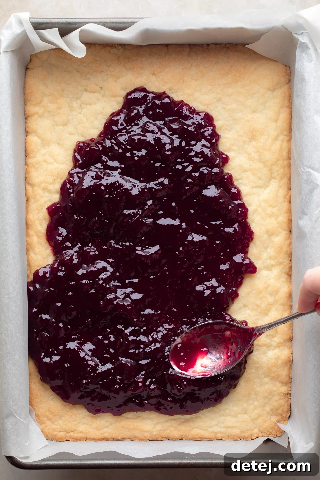 A spoon gently spreads a generous layer of vibrant red raspberry preserves evenly over a warm, golden-baked shortbread cookie crust in a baking dish, preparing the base for the next step.