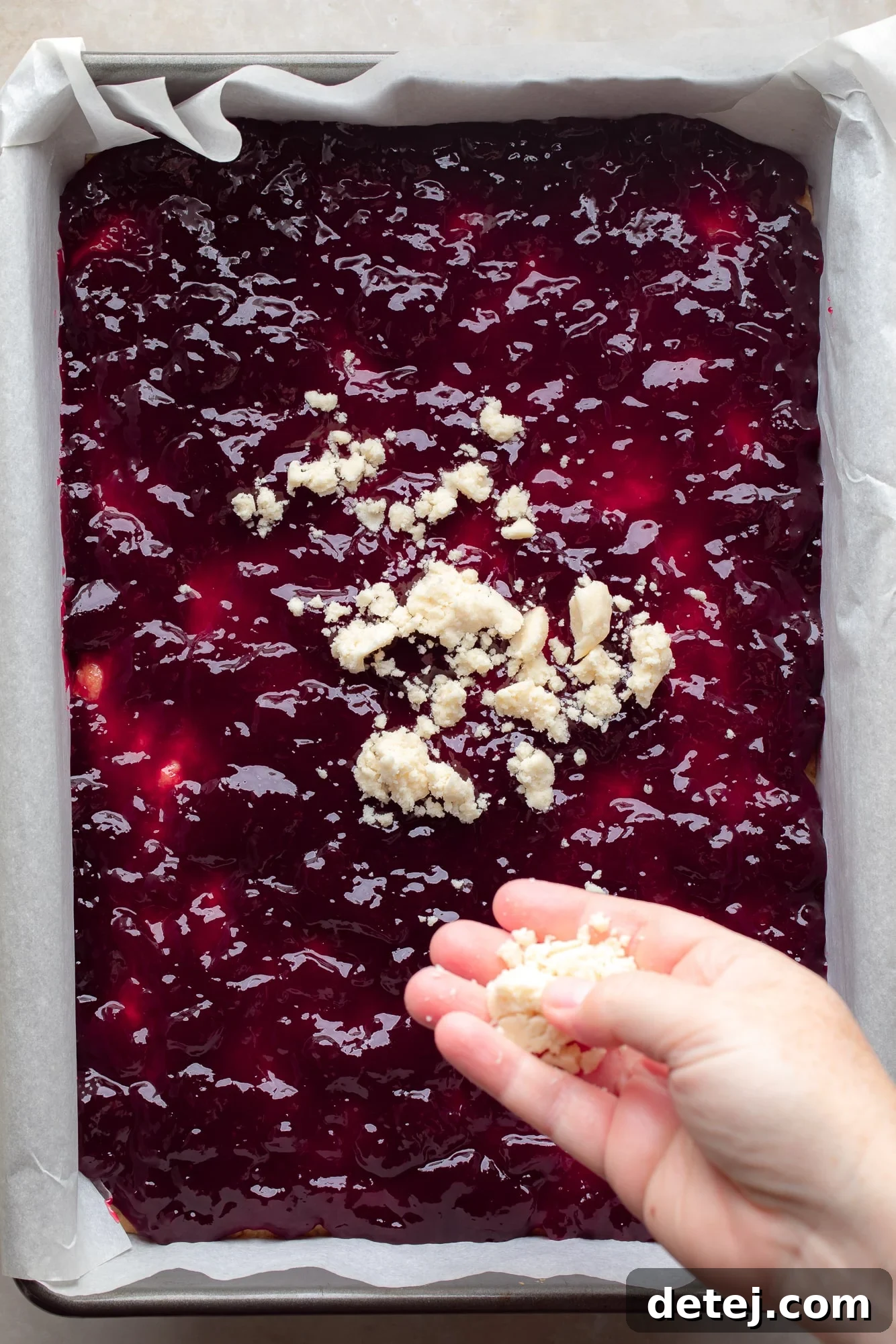 A woman's hand carefully sprinkles a delightful, crumbly shortbread topping over a bubbling layer of raspberry preserves, nestled atop a baked crust in a baking dish, creating the signature texture of vegan raspberry bars.