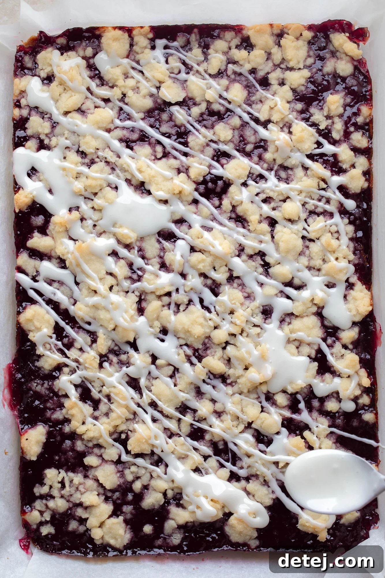 A spoon carefully drizzles a smooth, white glaze over freshly baked vegan raspberry bars cooling in a baking dish, adding a final touch of sweetness and visual appeal.