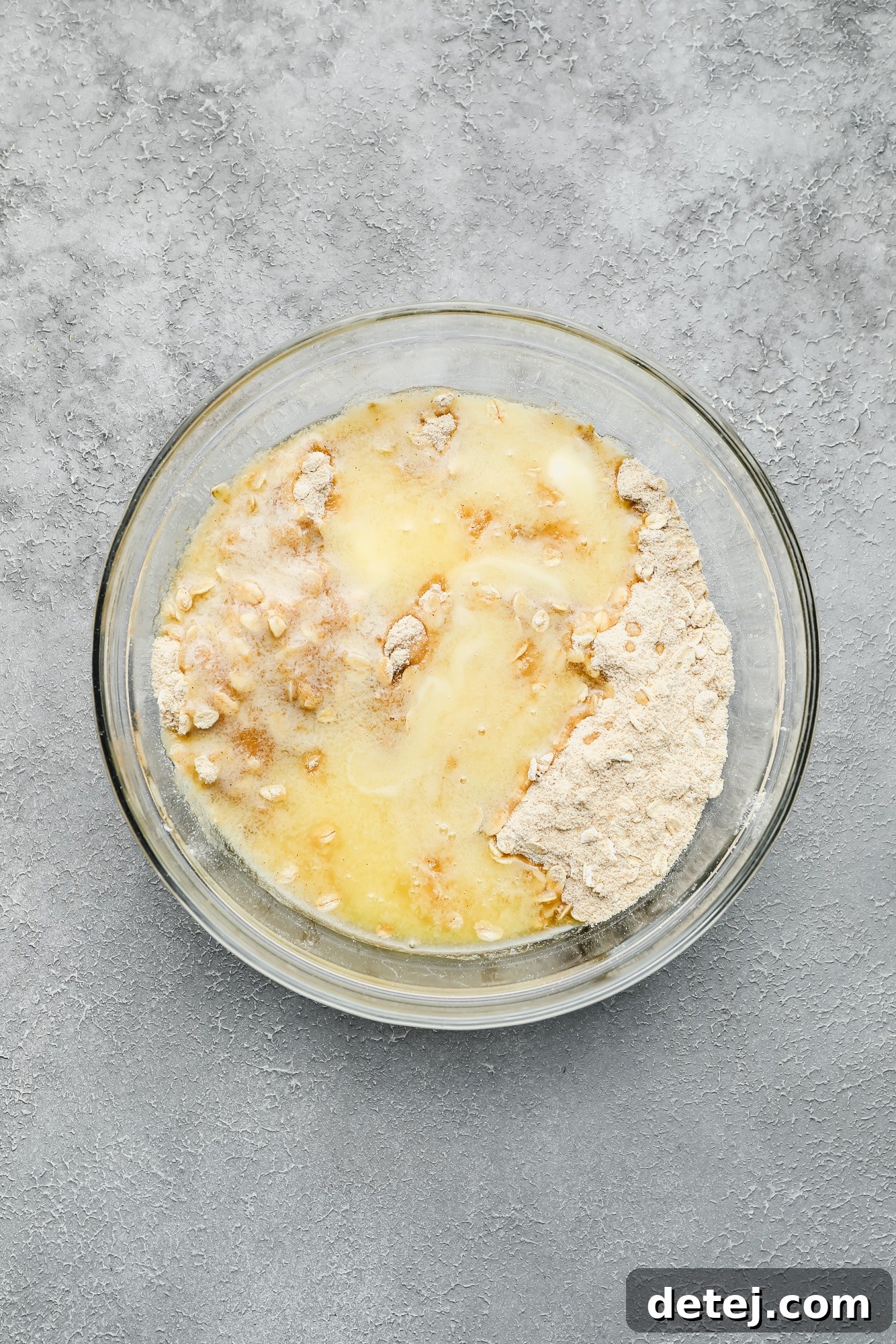 Melted vegan butter being poured over the mixture of flour, brown sugar, oats, and cinnamon in a glass bowl.