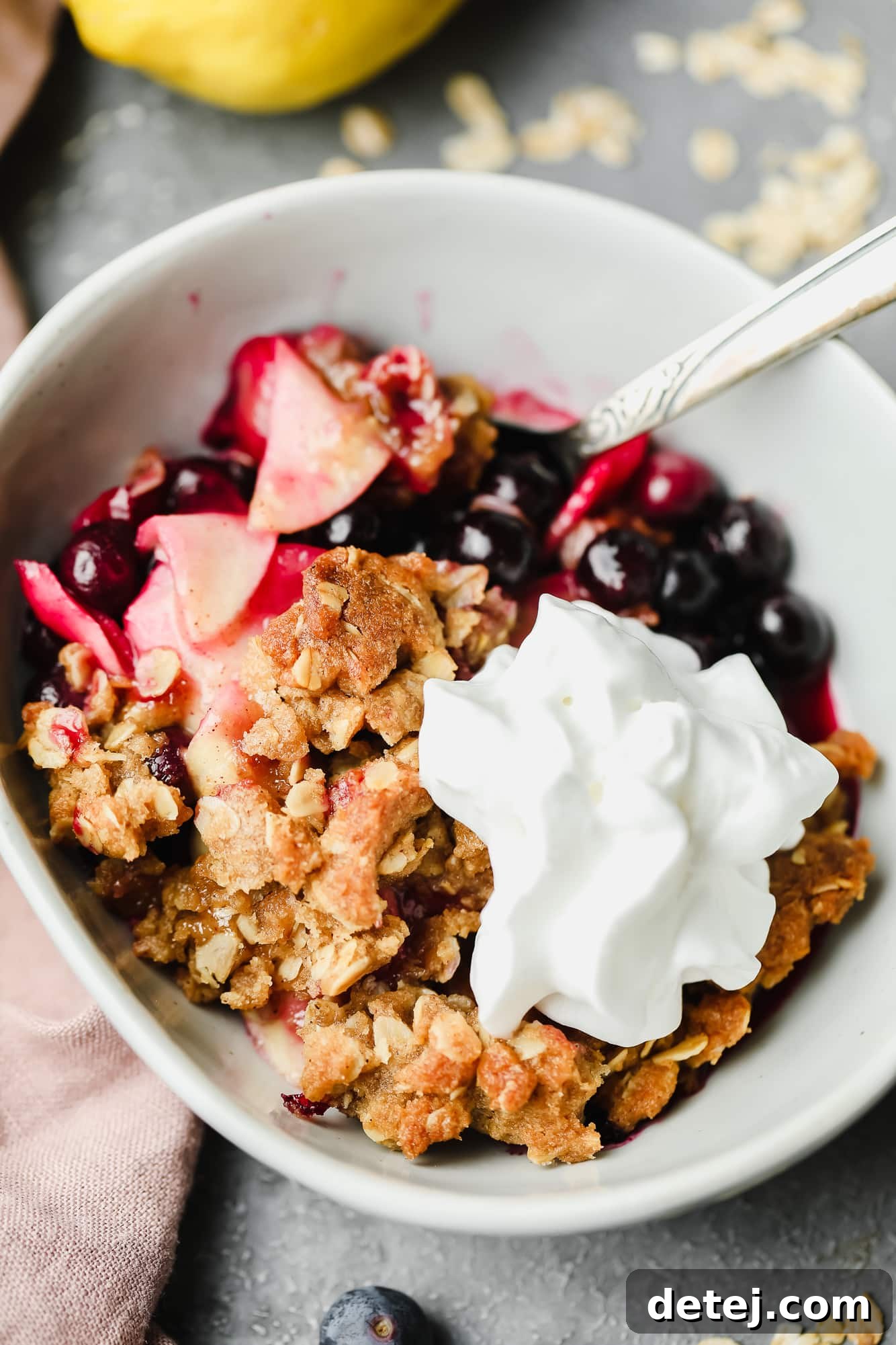 A close-up of a serving of baked apple and blueberry crumble, adorned with vegan whipped cream, glistening with fruit juices.