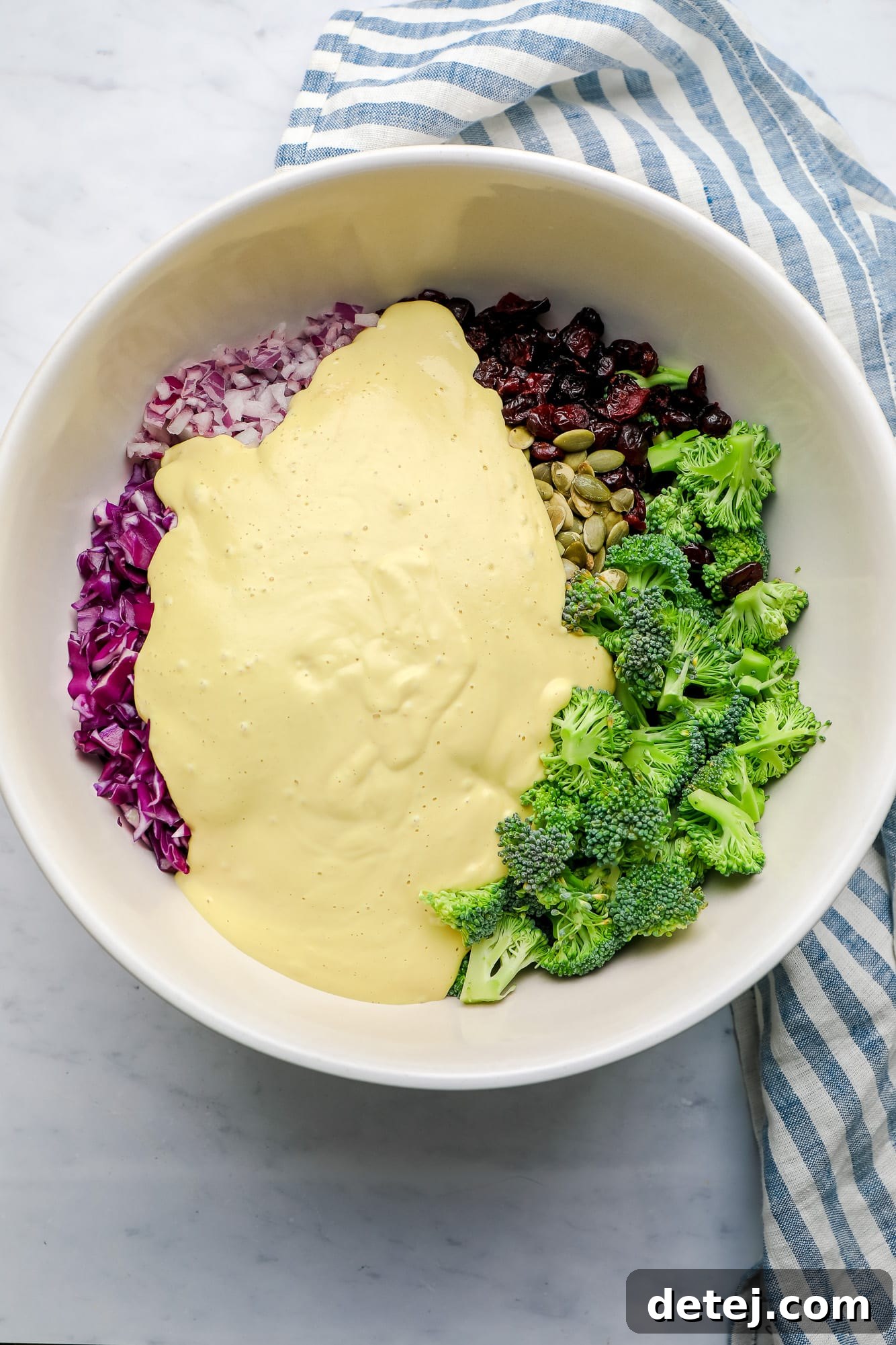 The vibrant ingredients for vegan broccoli salad, with the creamy yellow dressing poured over them, ready to be tossed in a large white bowl.
