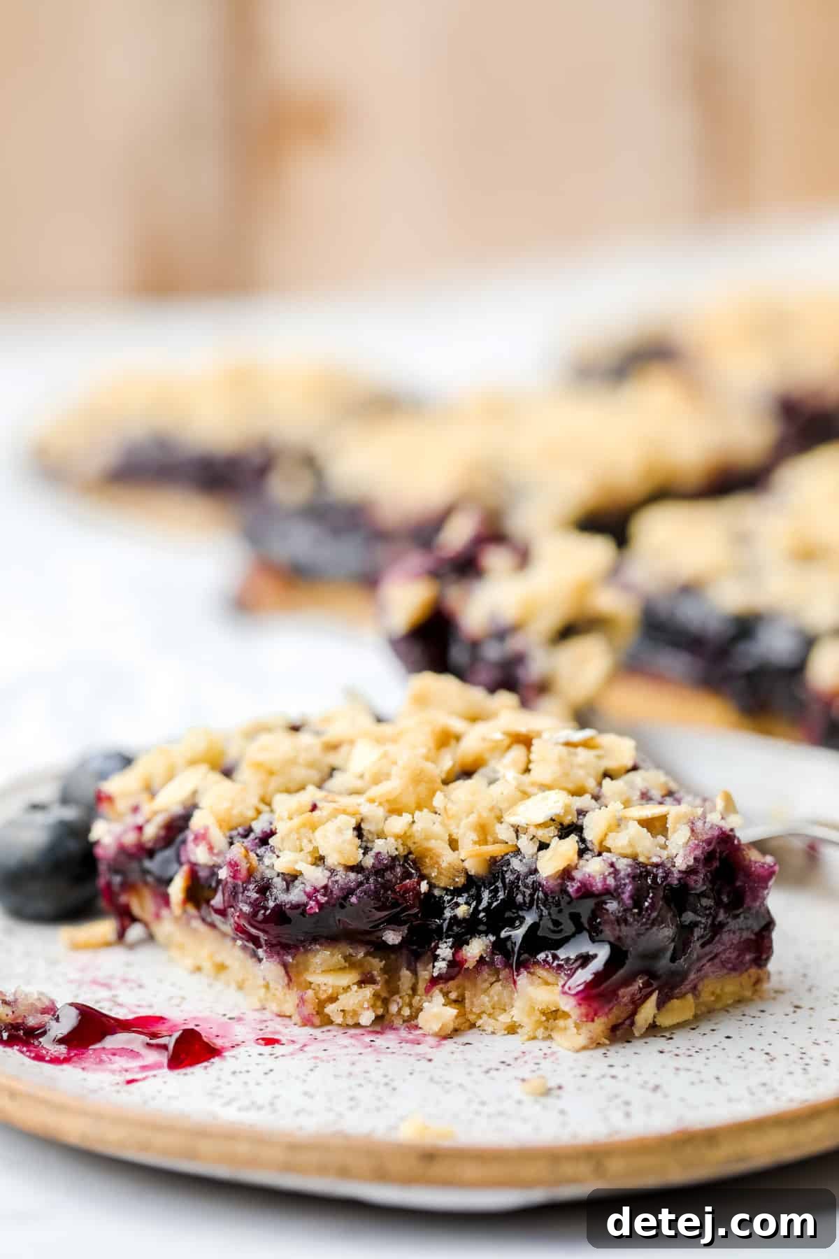 A beautifully speckled plate showcasing a single vegan blueberry pie bar with a bite taken out, revealing its generous blueberry filling and golden-brown oatmeal crumble.