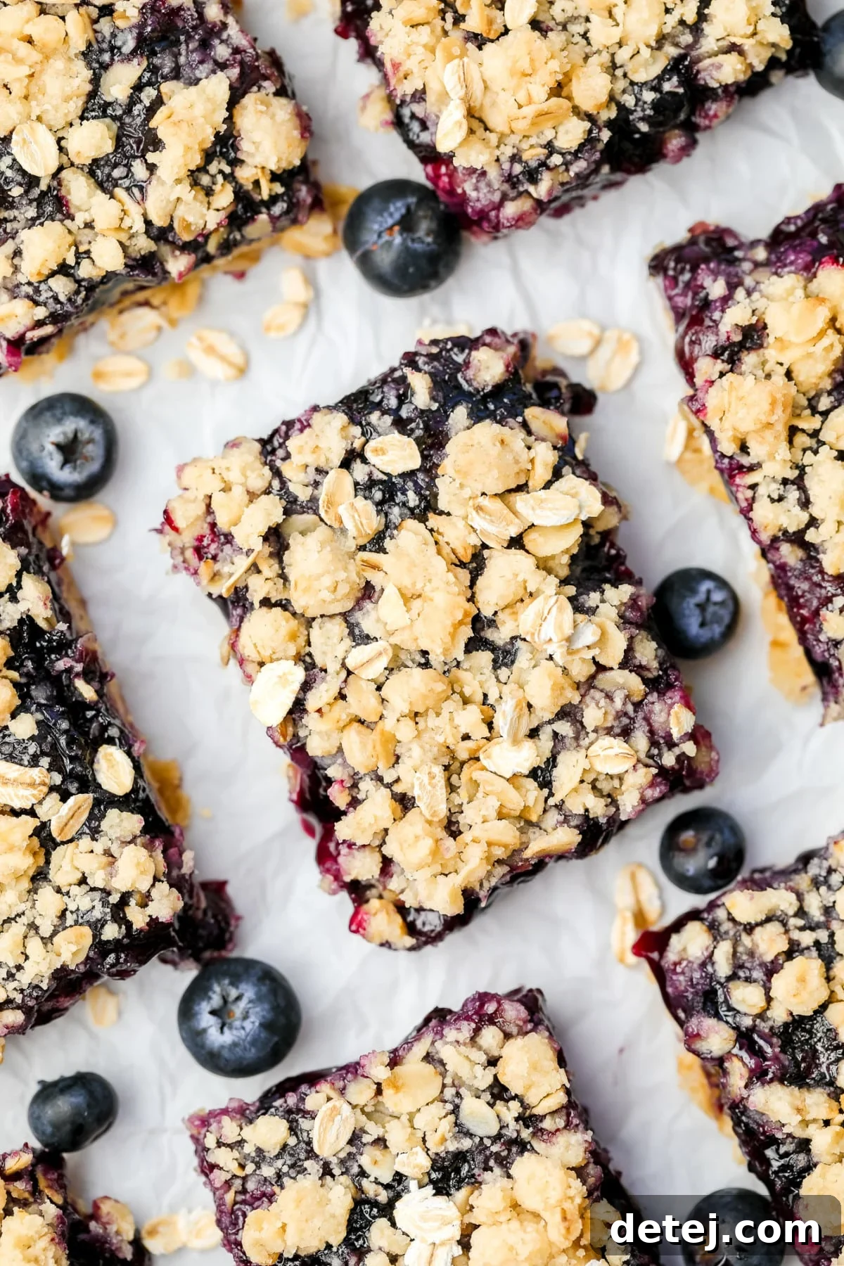 An overhead shot of freshly baked blueberry pie bars in a square pan, adorned with a golden-brown crumb topping, surrounded by fresh blueberries on a pristine white background.