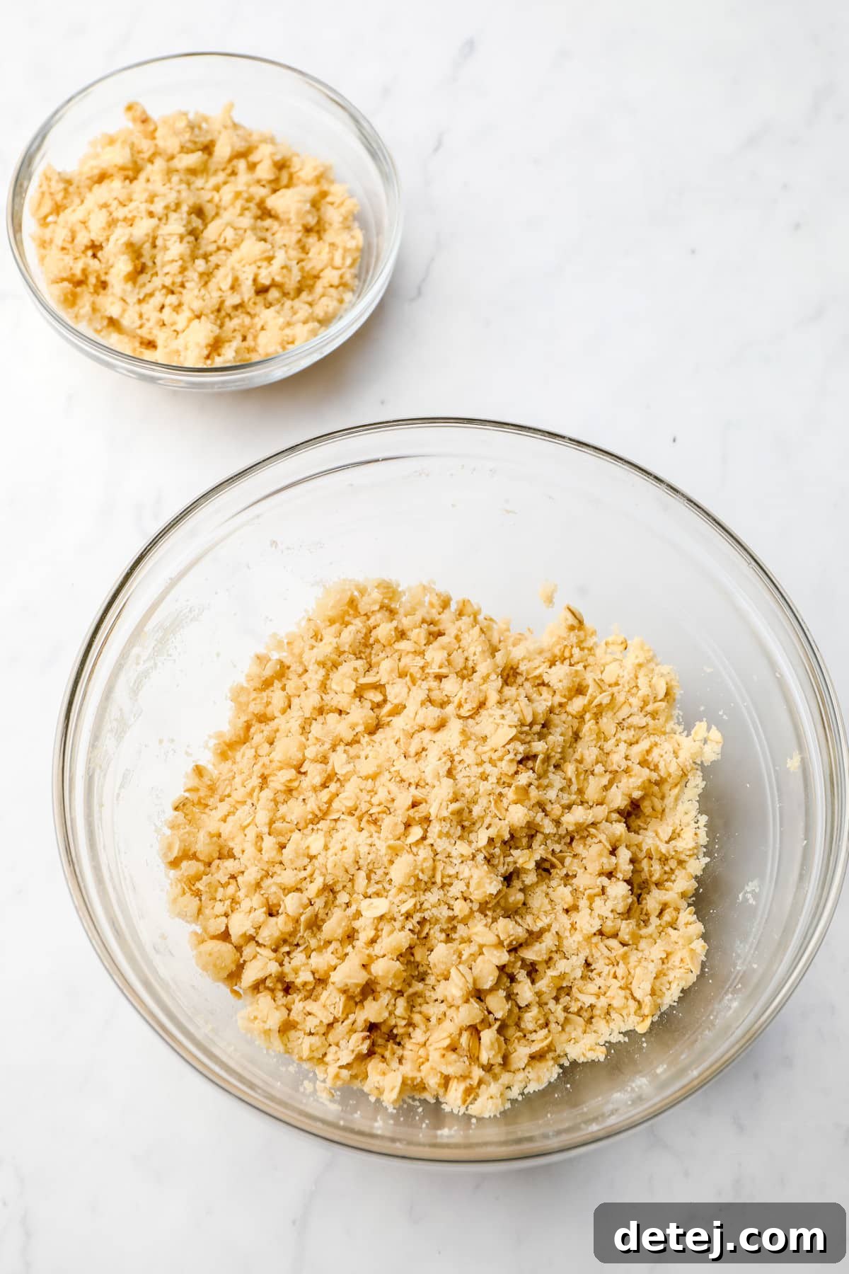 Two bowls on a countertop, one larger containing the bulk of the crumble mixture, and a smaller bowl holding the reserved portion for the topping.