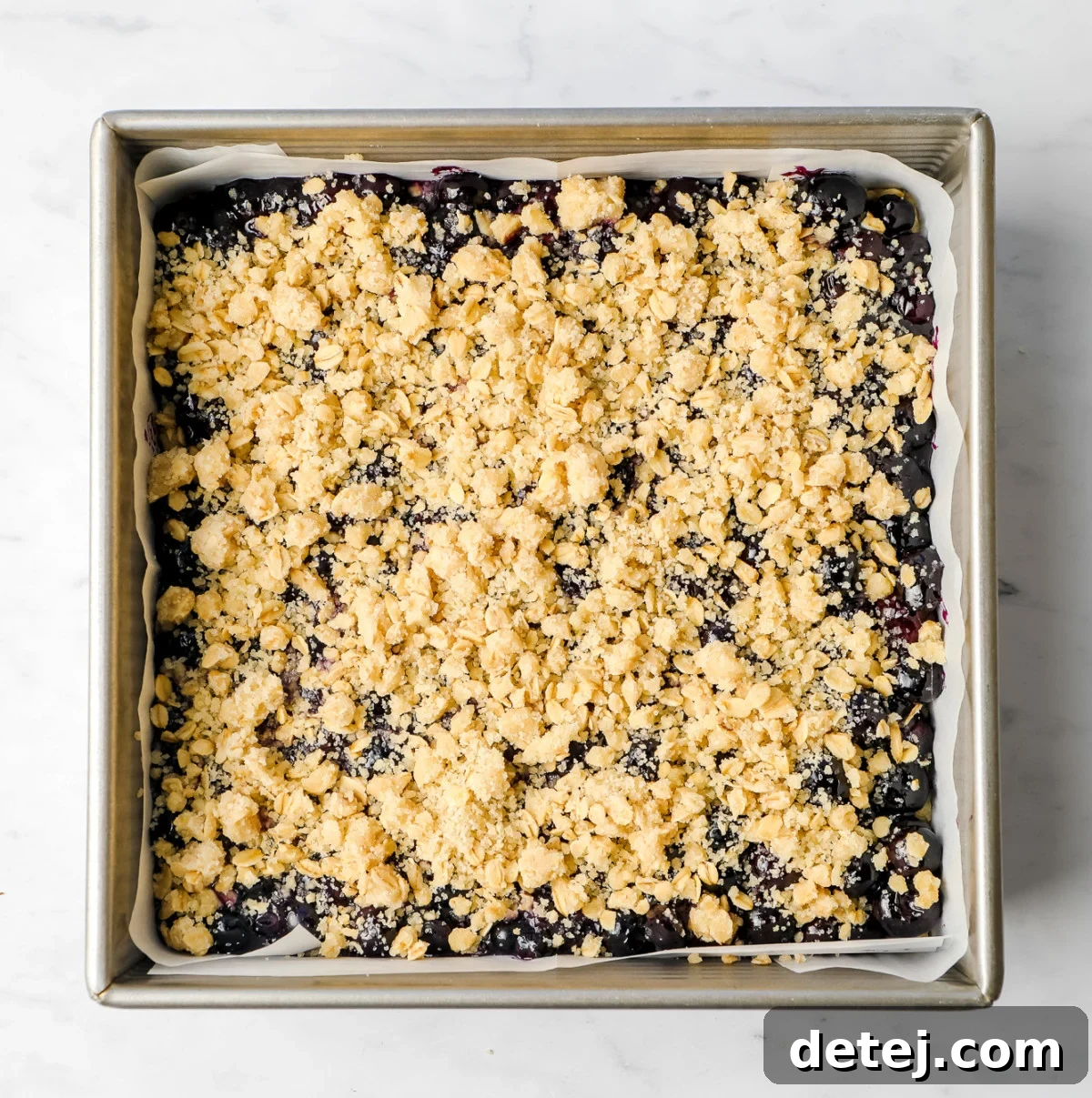 An uncooked square pan containing the blueberry mixture covered with a layer of the crumb topping, prepared for baking.