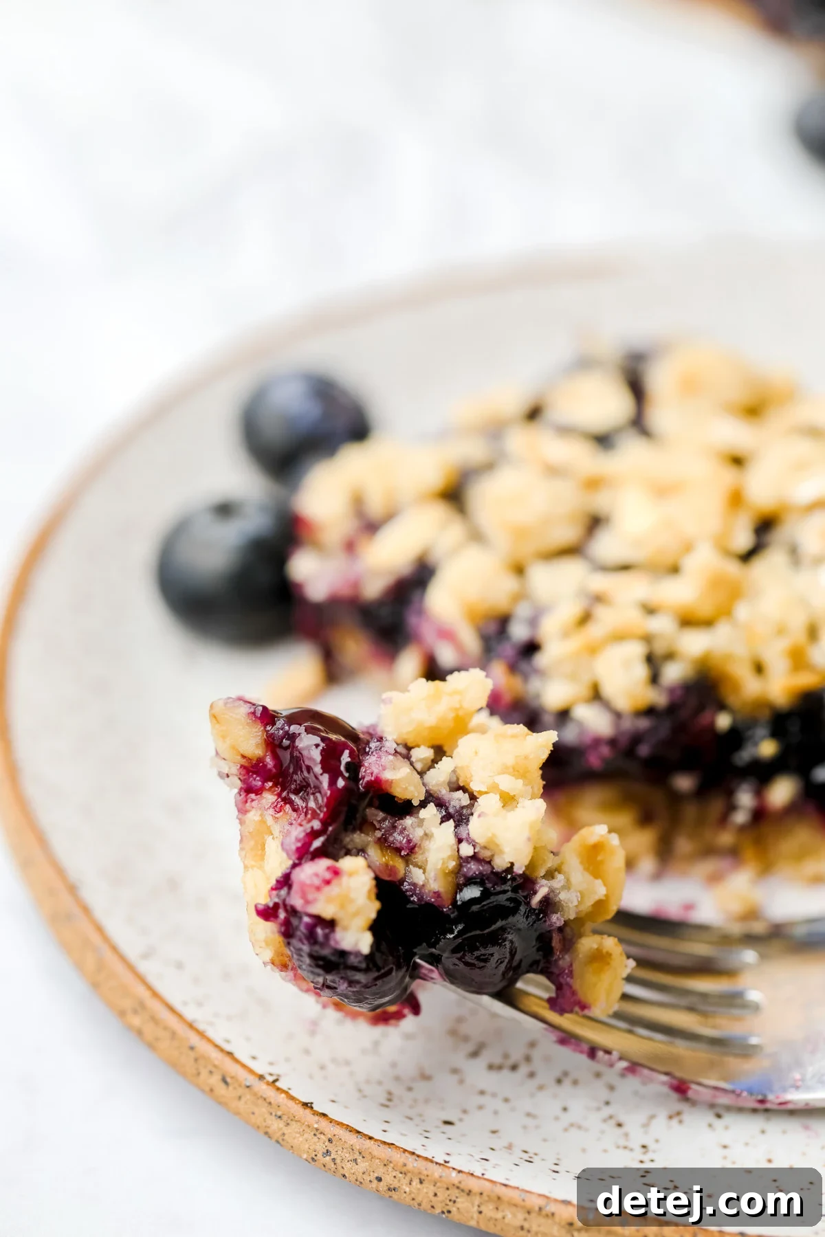 A fork holding a bite of a vegan blueberry pie bar, clearly showing the cooked blueberry filling and the crunchy crumb topping.