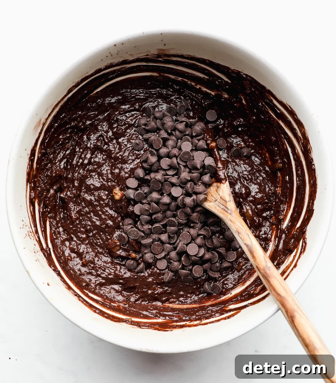 using a wooden spoon to stir chocolate chips into chocolate batter in a white bowl, showing the rich, dark mixture.