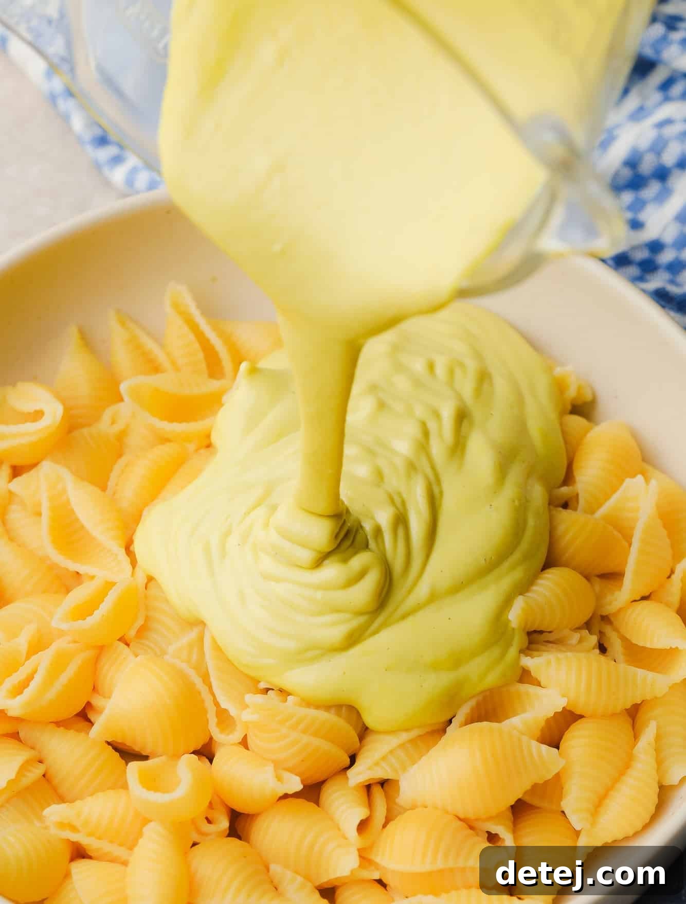 A generous stream of golden cashew cheese sauce cascades over a large bowl of cooked pasta, promising a creamy and flavorful meal.