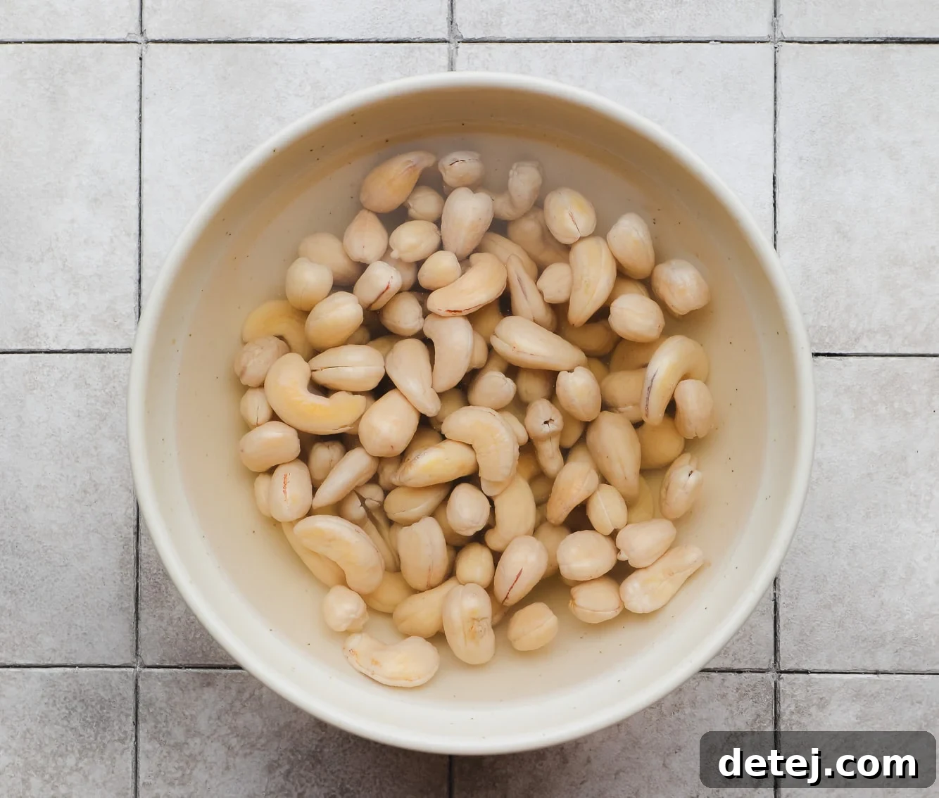 Raw cashews soaking in a beige bowl, submerged in water, preparing them for blending into a creamy sauce.