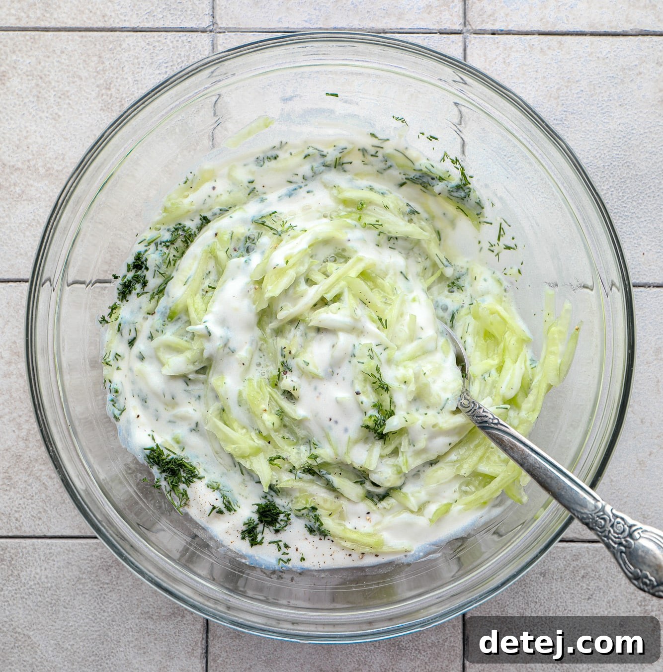 A glass bowl containing the vibrant ingredients for homemade tzatziki sauce, being gently stirred to combine the refreshing flavors.