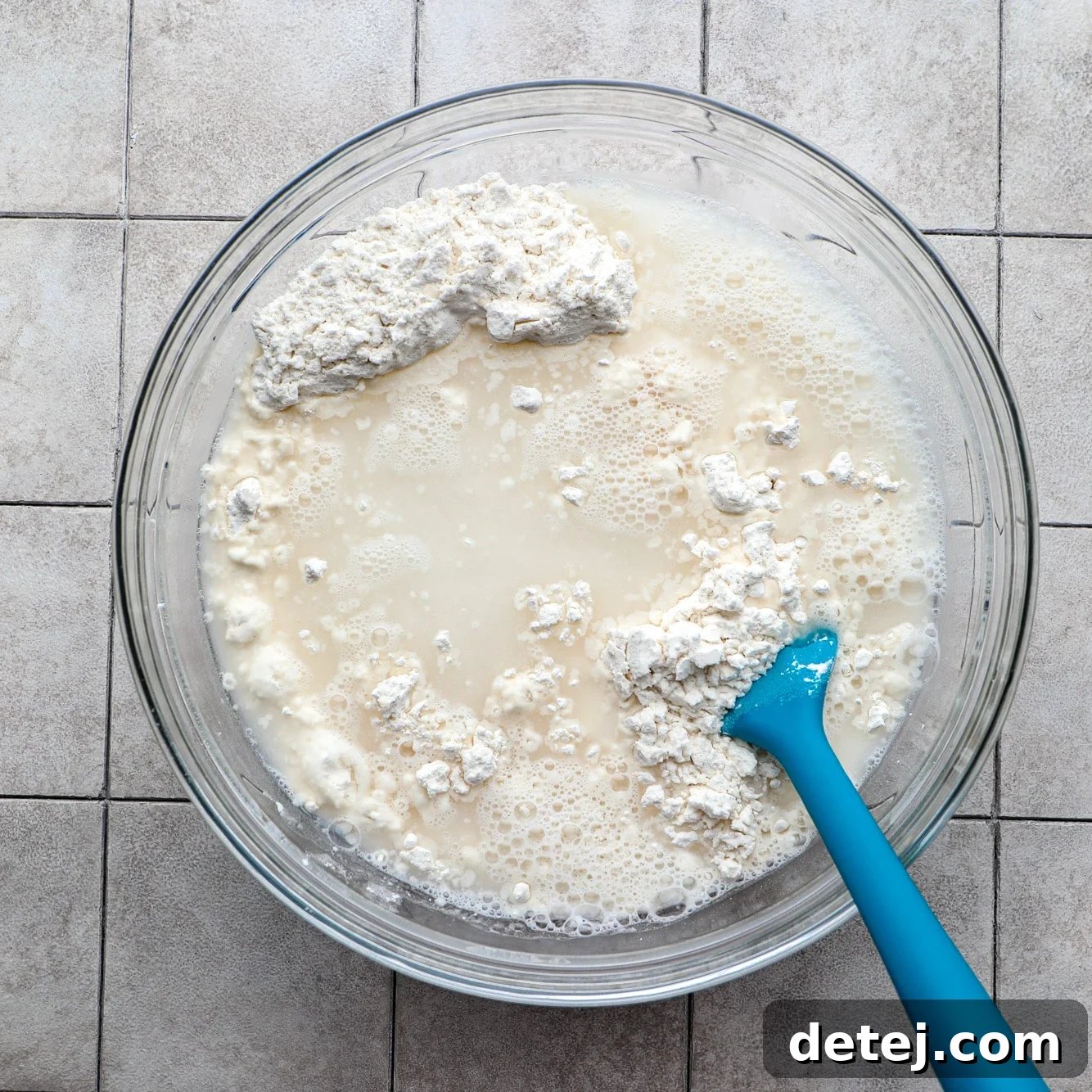 A blue spatula stirring together the focaccia dough ingredients in a glass bowl until just combined.