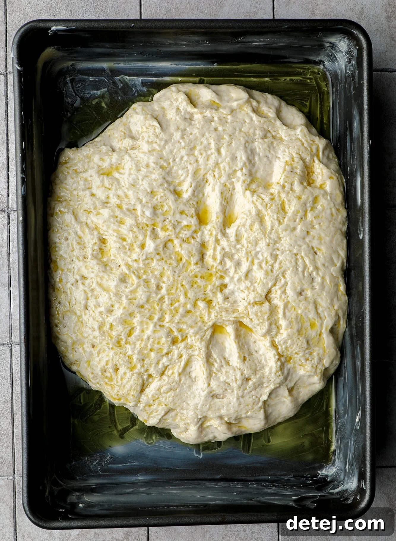 Oiled focaccia dough resting in a large black baking pan before the second rise.
