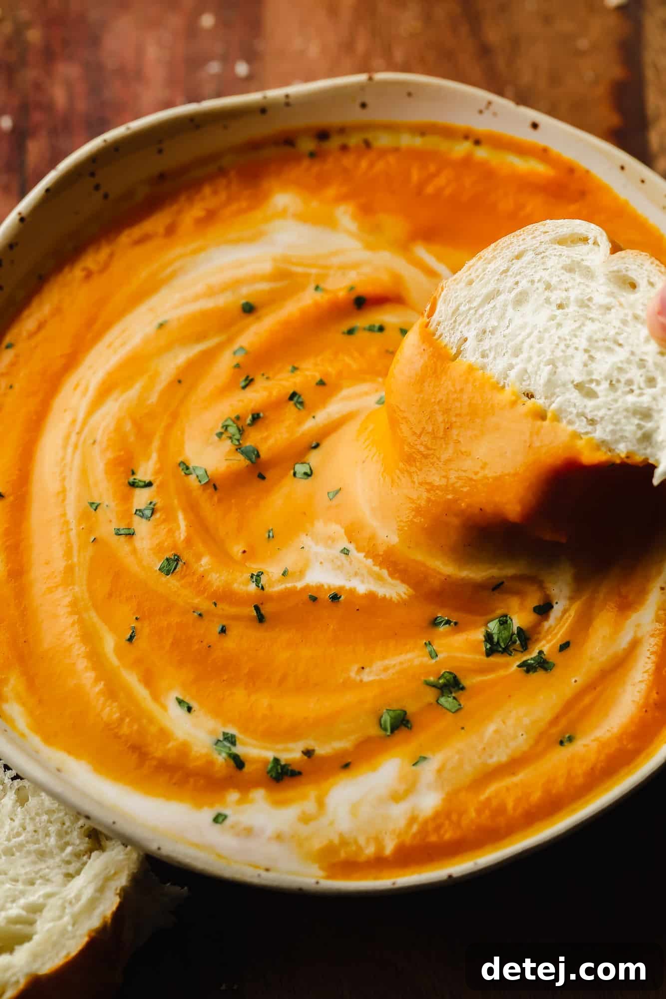 A hand dipping a piece of crusty bread into a steaming bowl of creamy vegan pumpkin soup, garnished with a swirl of coconut milk and fresh herbs.